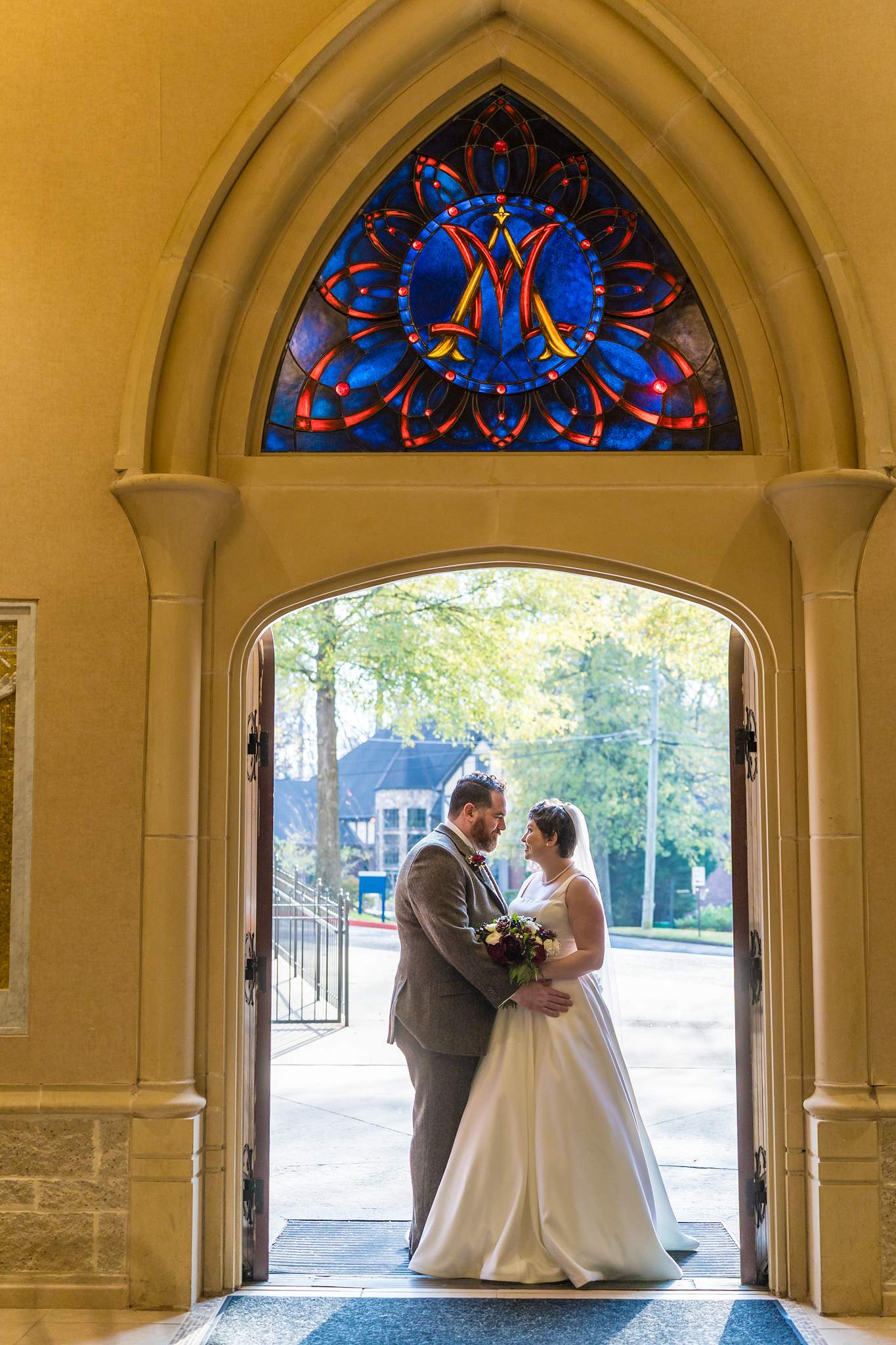 Couple in the arched church doorway under a stained glass window at Our Lady of the Assumption