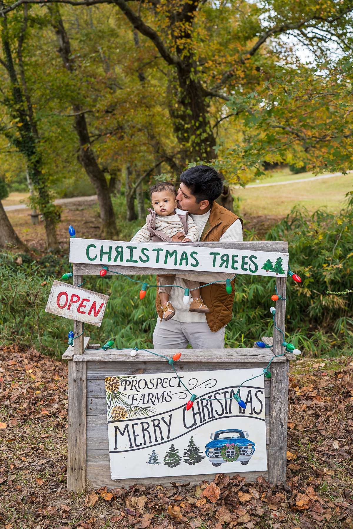 Family with their baby behind the Christmas tree stand at Prospect Farms