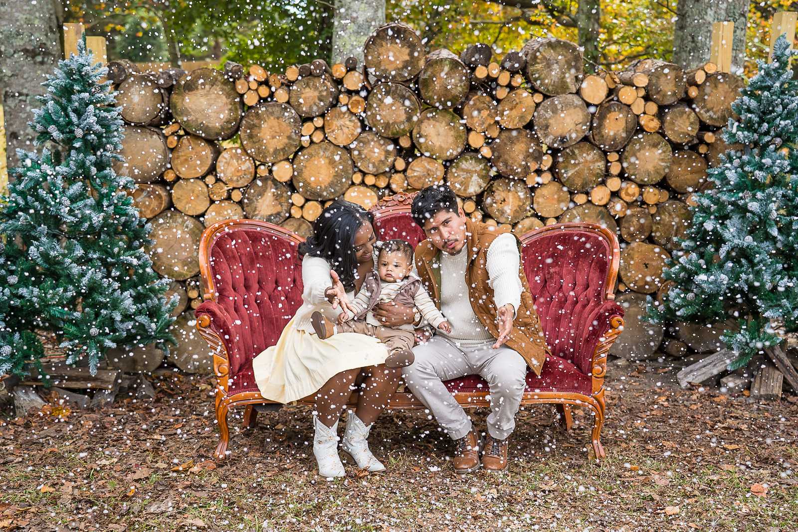 Parents and baby on the red velvet couch in front of the Log Wall as snow falls at Prospect Farms