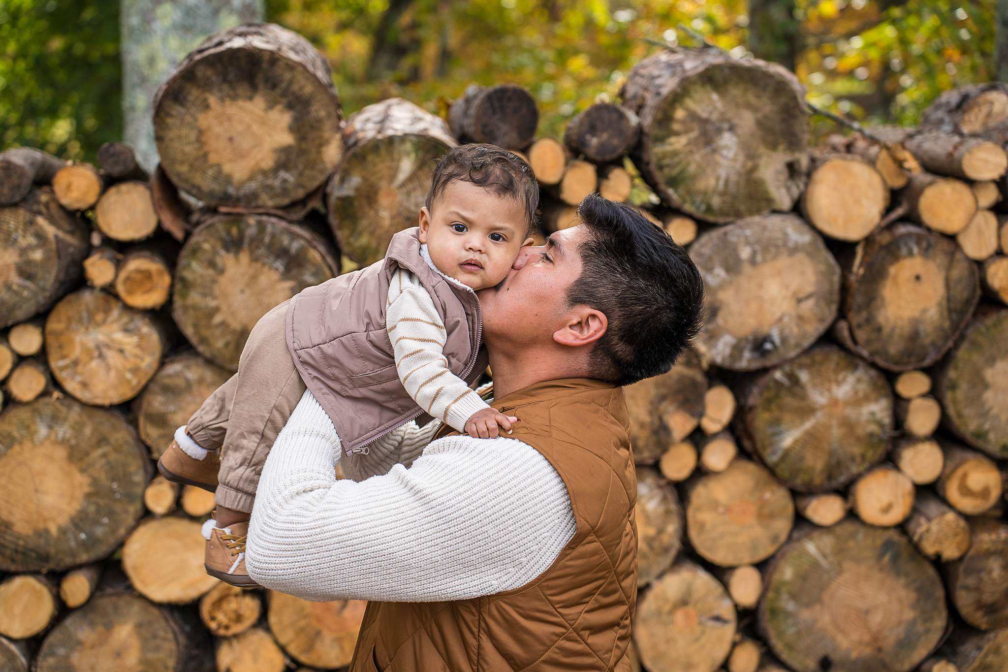 Dad lifting his baby as snow falls in front of the Log Wall at Prospect Farms