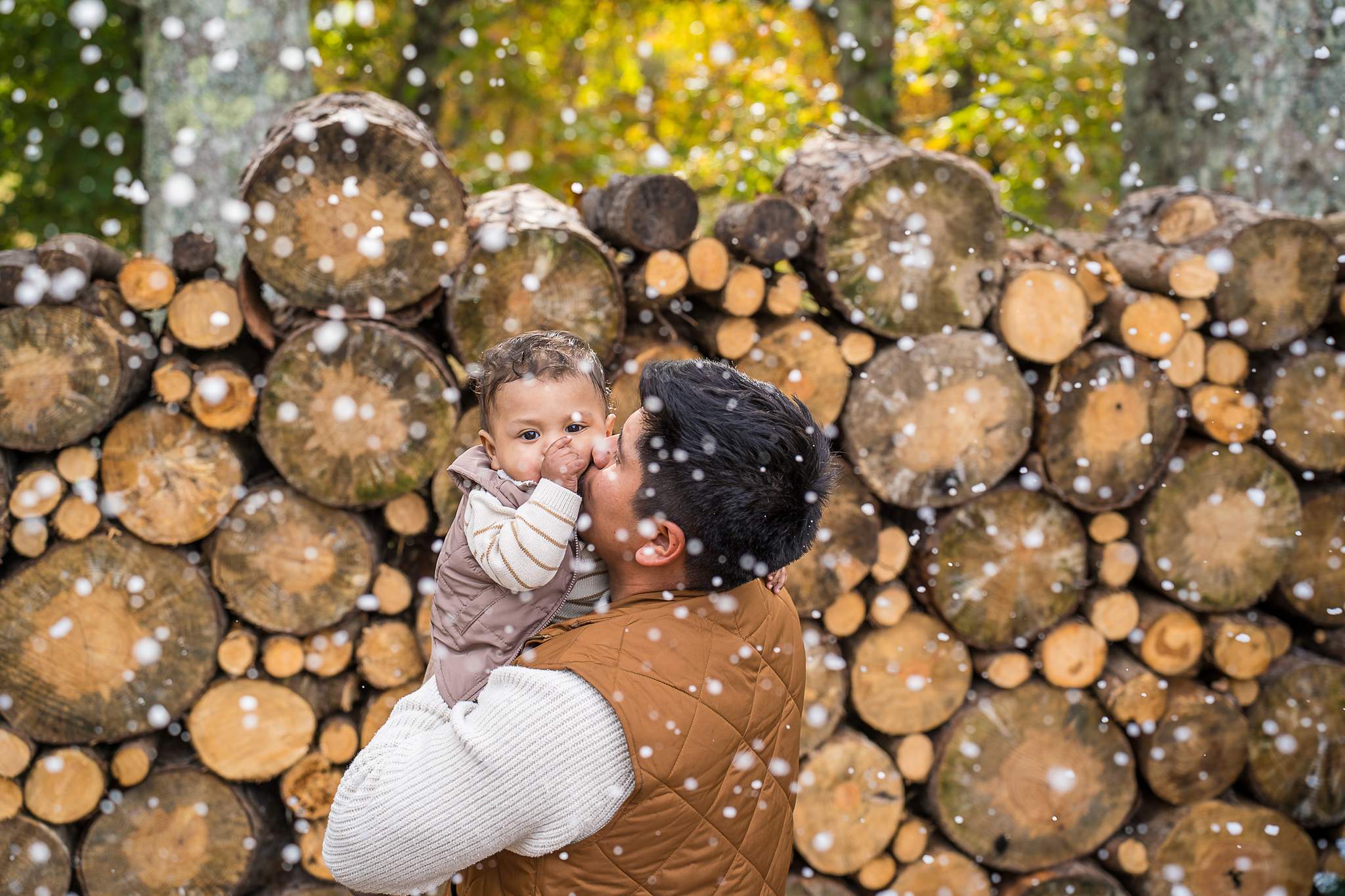 Dad kissing his baby in front of the Log Wall with falling snow at Prospect Farms