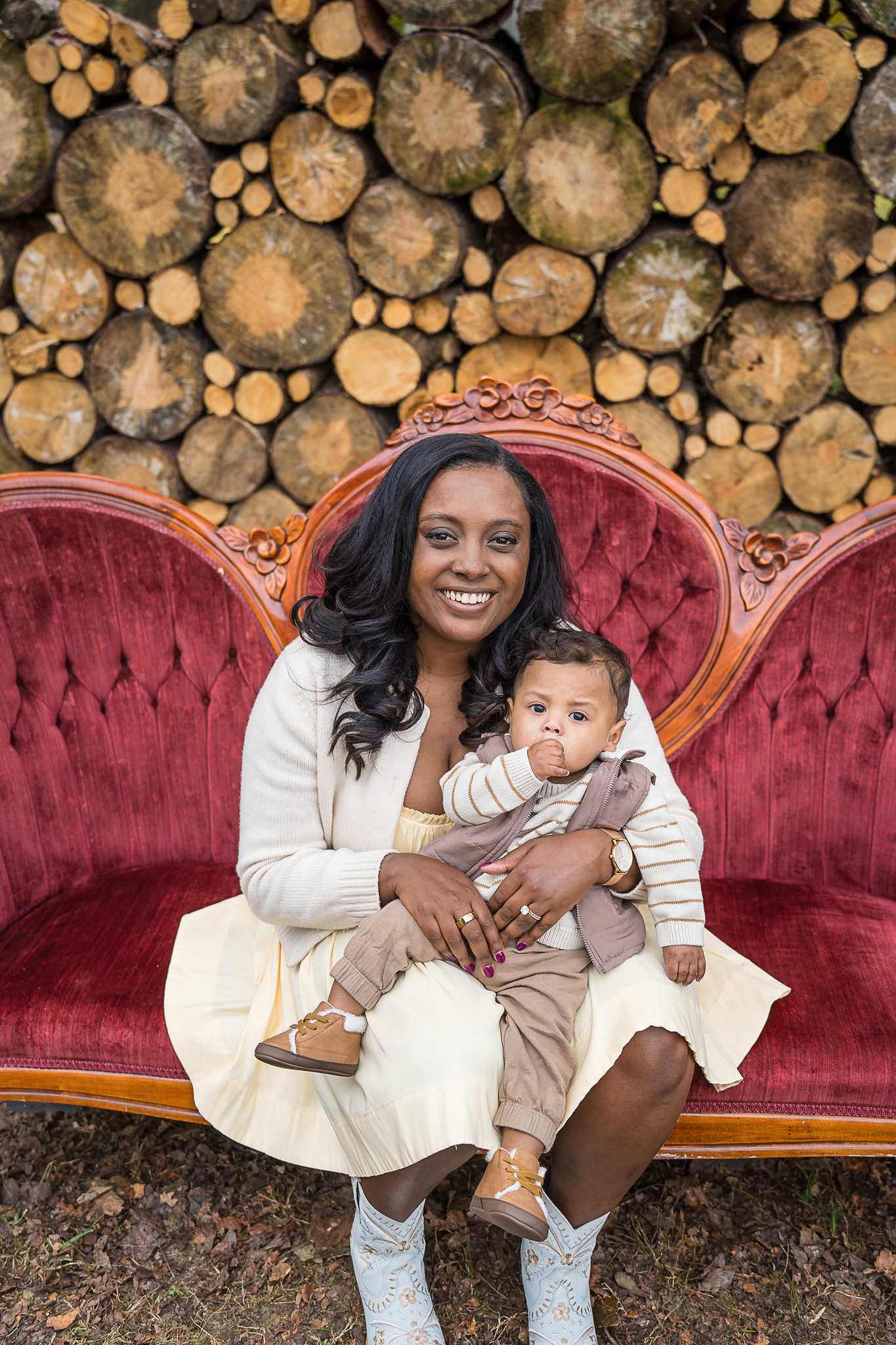 Mother in cream holding her baby on the red velvet couch at the Log Wall set