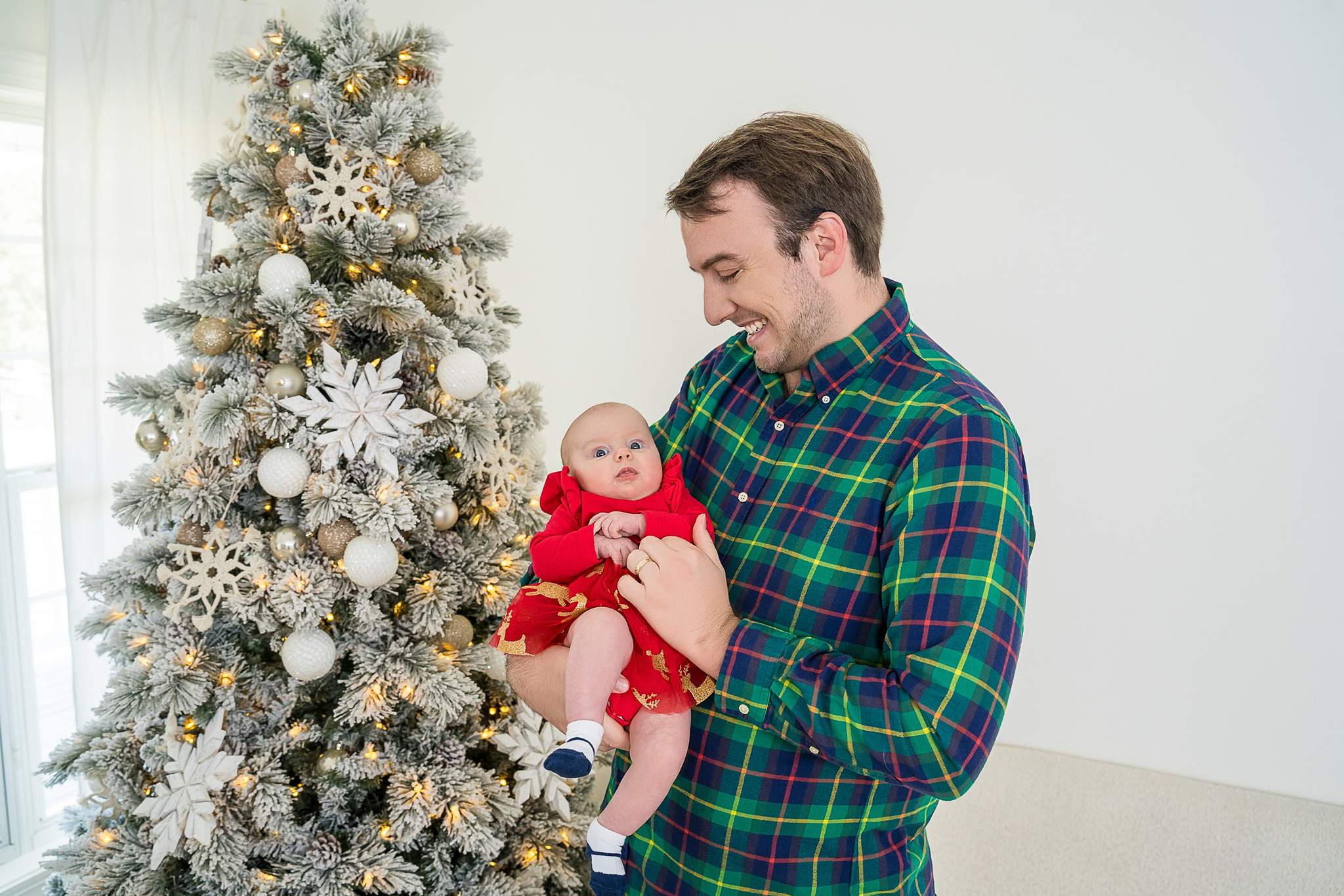 Dad in a green plaid shirt holding his baby in a red dress beside the flocked Christmas tree in the Lounge