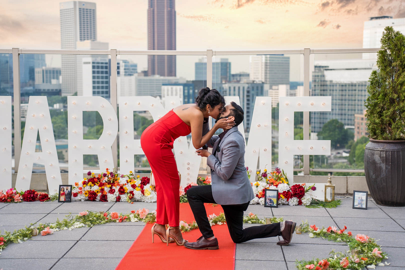 Couple kissing moments after a rooftop proposal, with Marry Me letters, roses, and the Atlanta skyline behind them
