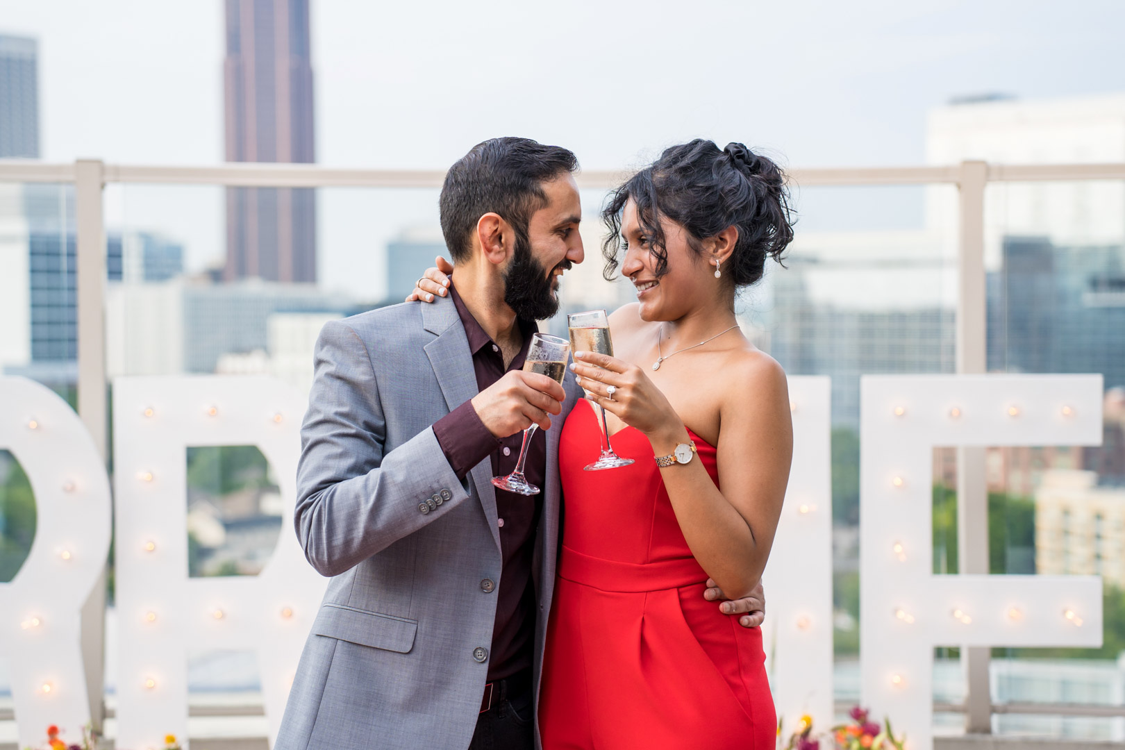 Newly engaged couple toasting with champagne beside lit Marry Me marquee letters on an Atlanta rooftop