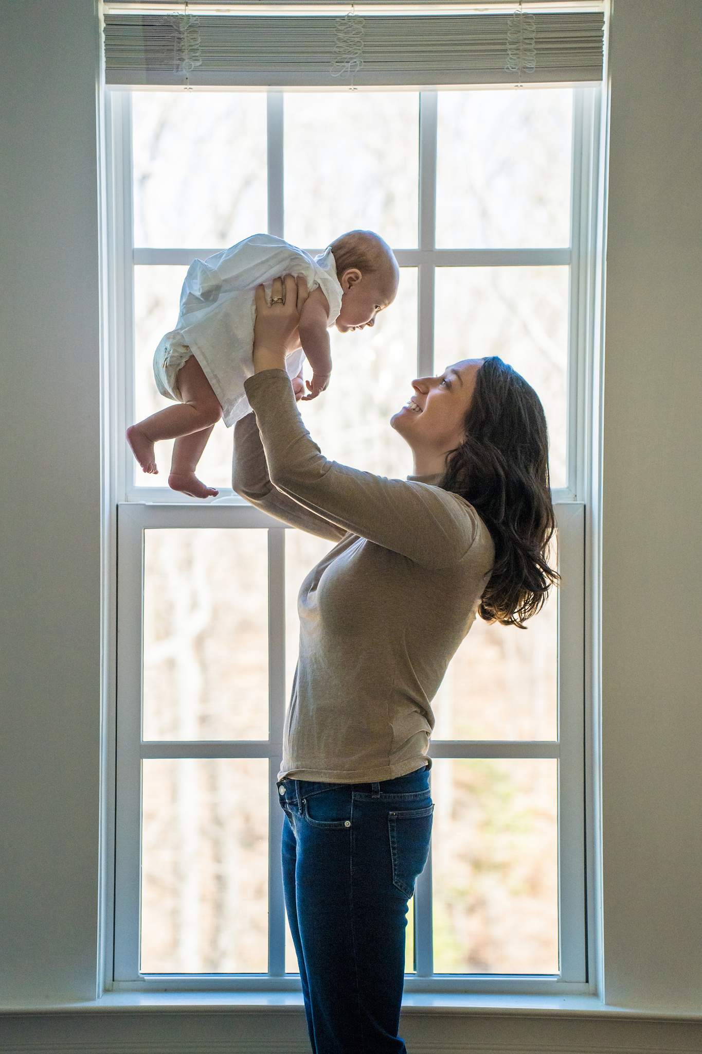 Mom in a camel ribbed sweater and dark jeans lifting her baby in white by a tall window
