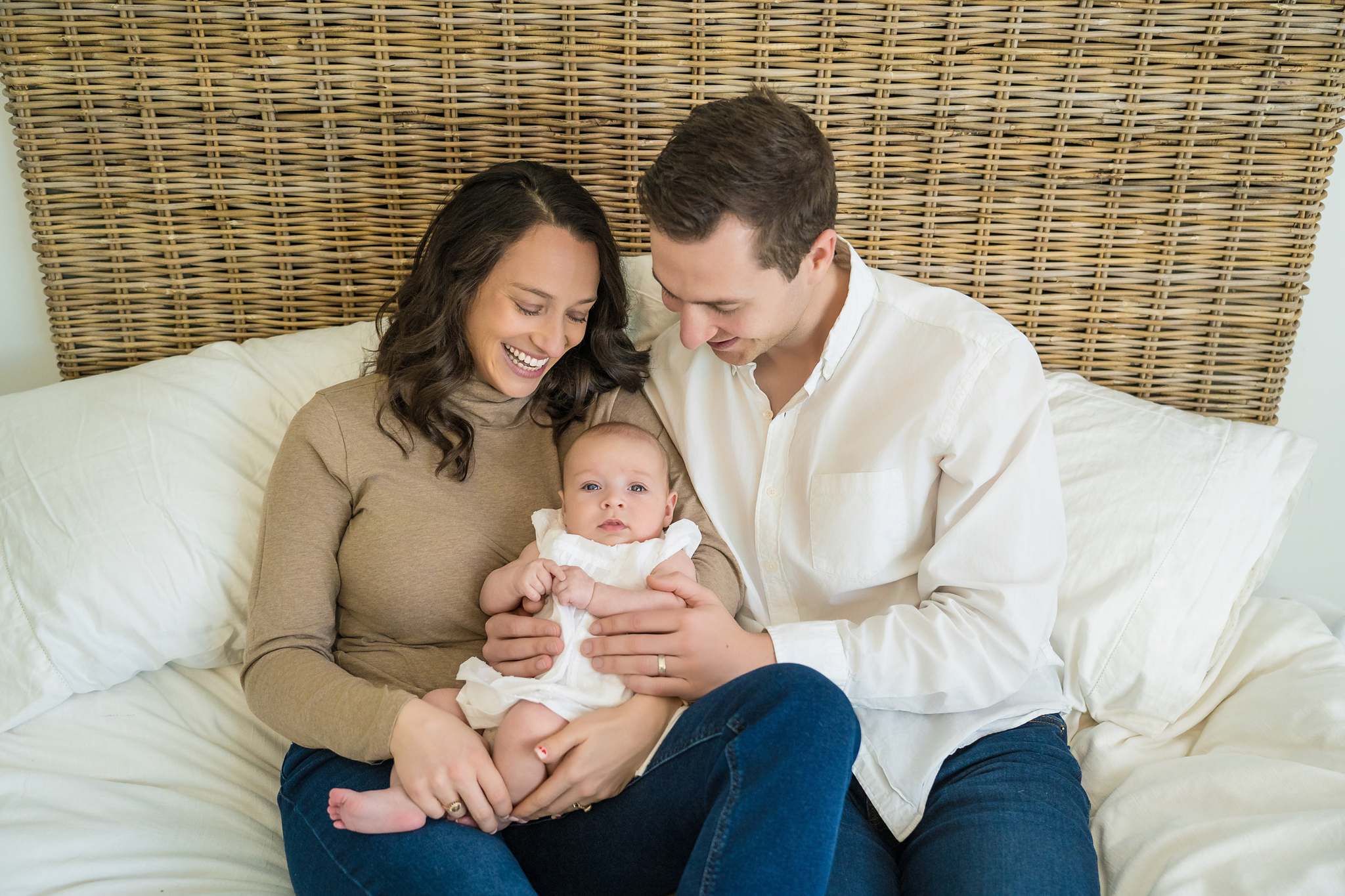 Neutral family outfits on a white bed: camel turtleneck, cream button-down, dark jeans and a baby in white