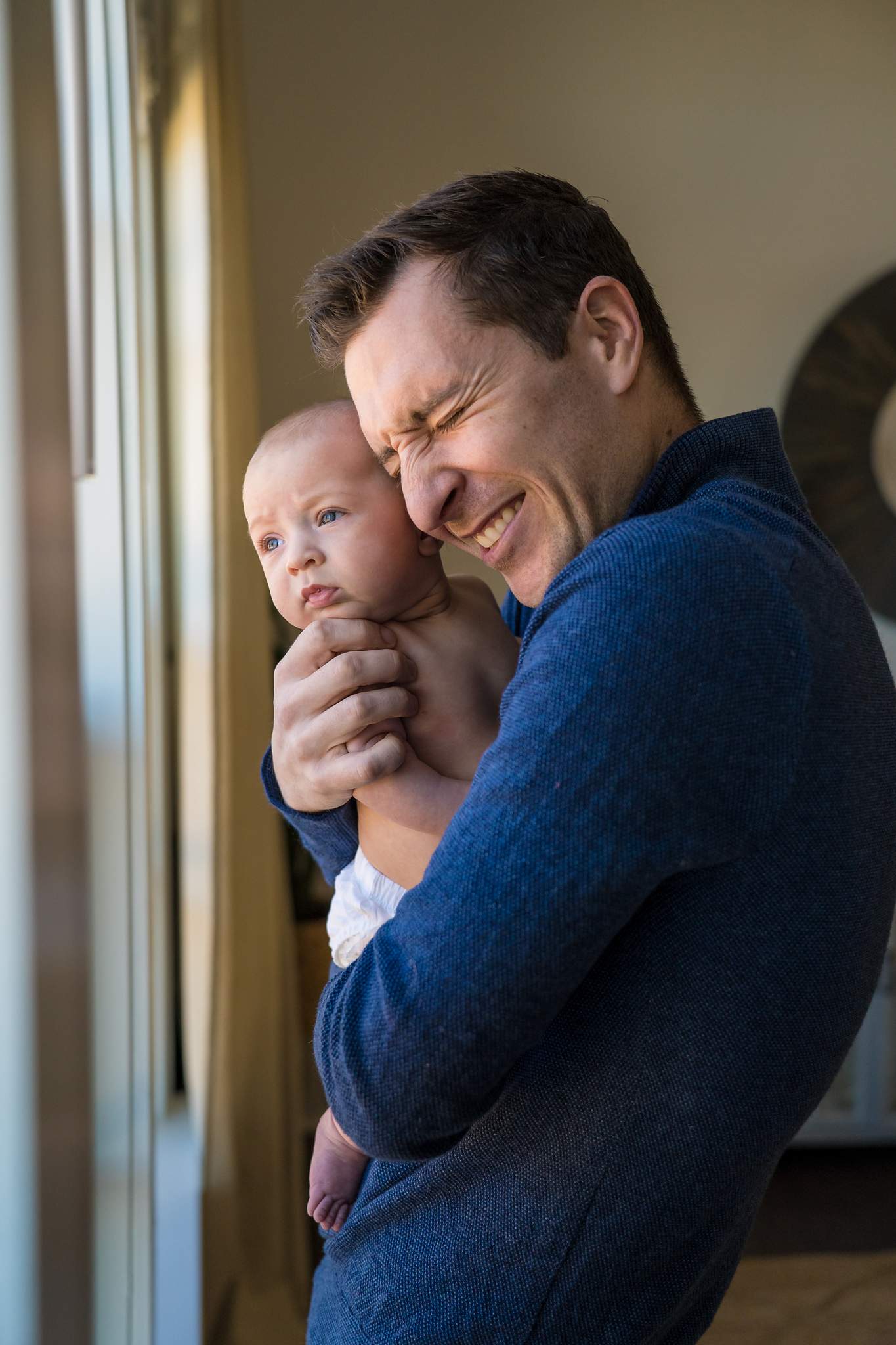 Dad in a navy quarter-zip sweater laughing as he holds his baby close by a window