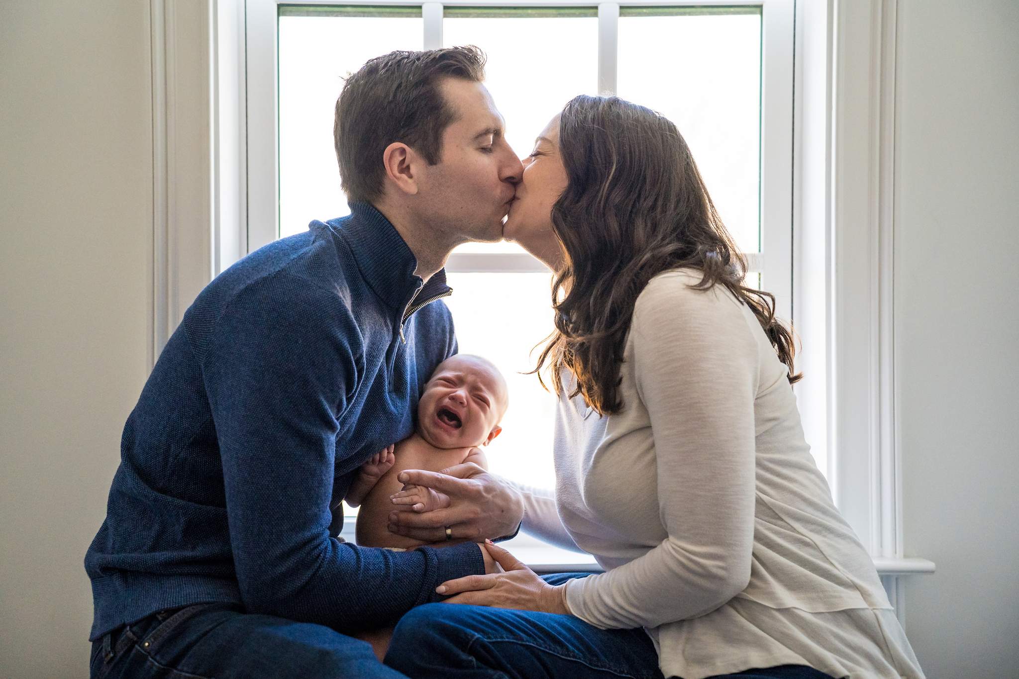Parents in a navy quarter-zip and oatmeal cardigan kissing over their fussy baby by a bright window