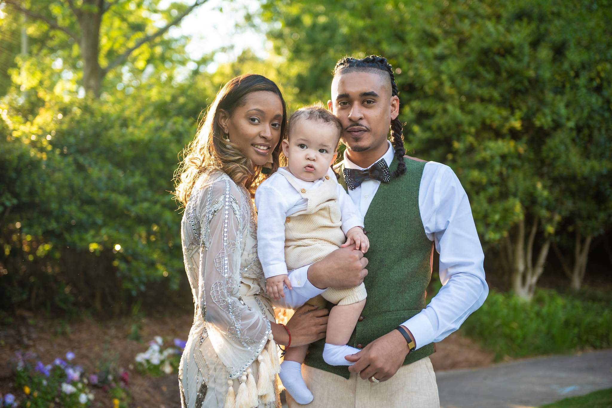 Cream beaded gown and olive green vest on parents holding their baby in cream in a sunlit park