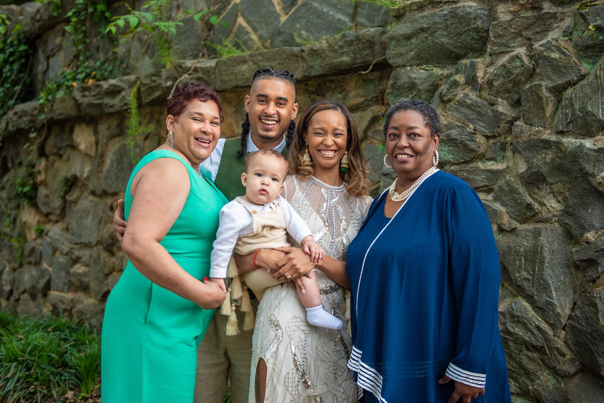 Relatives in teal and navy beside parents in cream and olive holding their baby by a stone wall