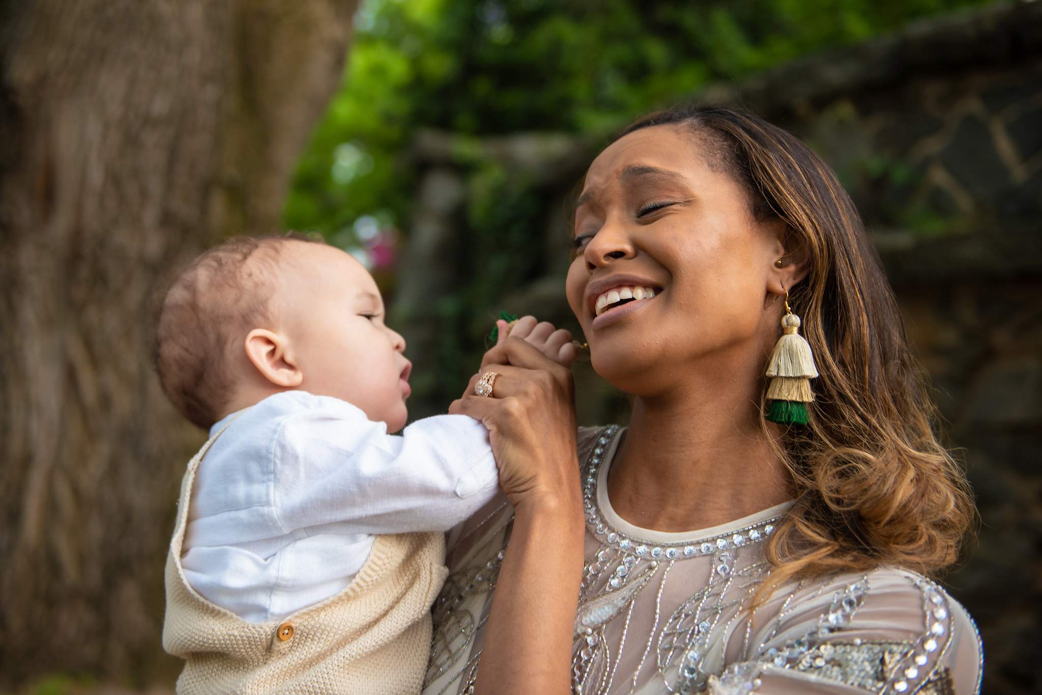 Mom in a beaded cream gown and green tassel earrings laughing with her baby in a white shirt