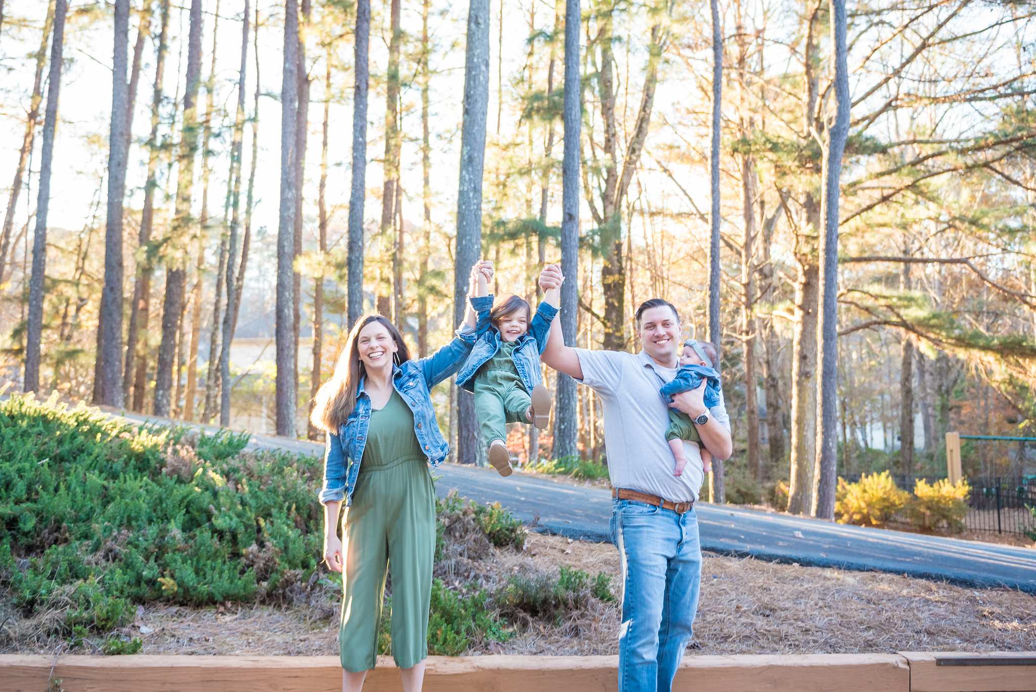 Parents in an olive jumpsuit and gray polo swinging a toddler in denim and olive between them