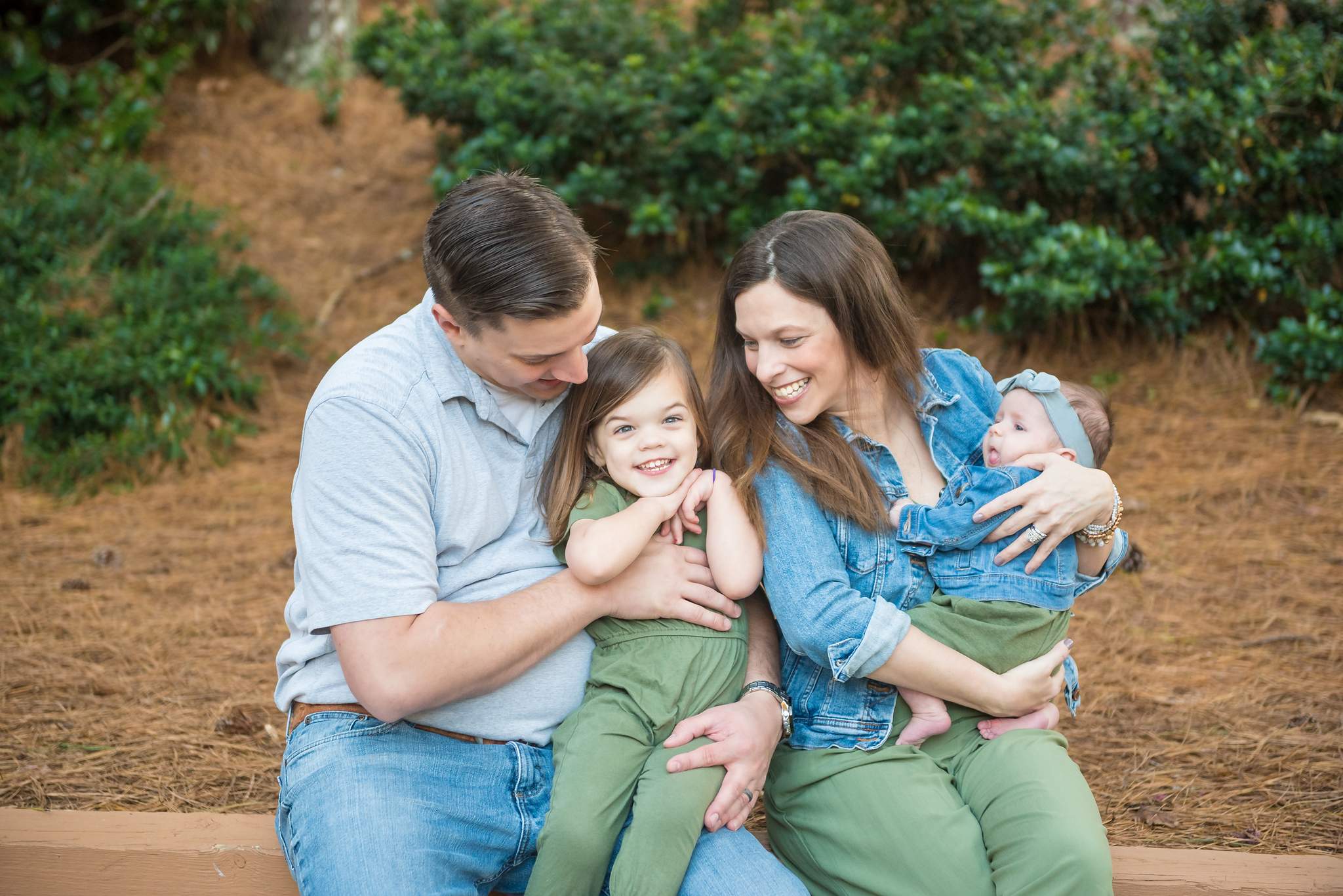 Family laughing on a wooden ledge in an olive jumpsuit, denim jackets, gray polo and jeans