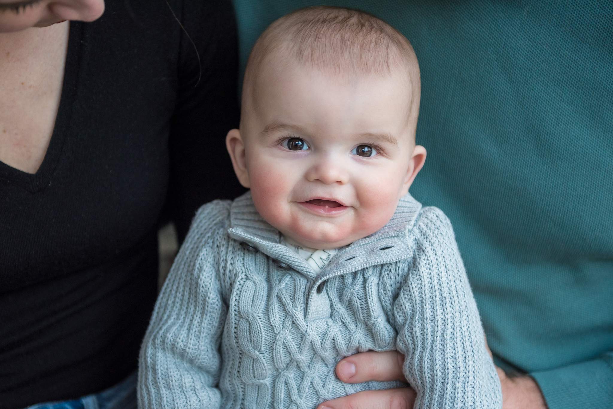 Baby in a soft gray cable-knit sweater held between parents in black and teal