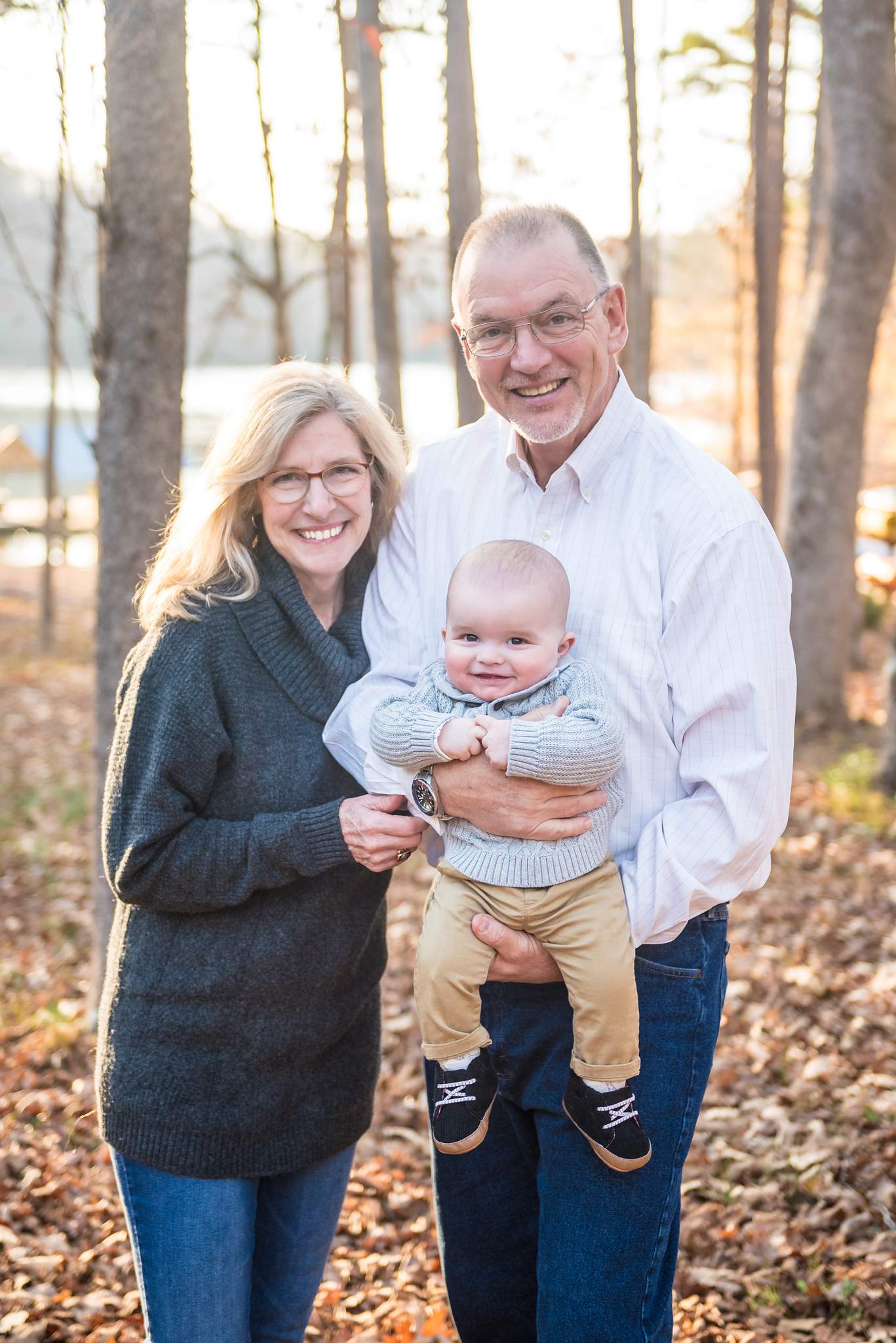 Grandparents in a charcoal sweater, white button-down and jeans holding a baby in gray knit among fall leaves