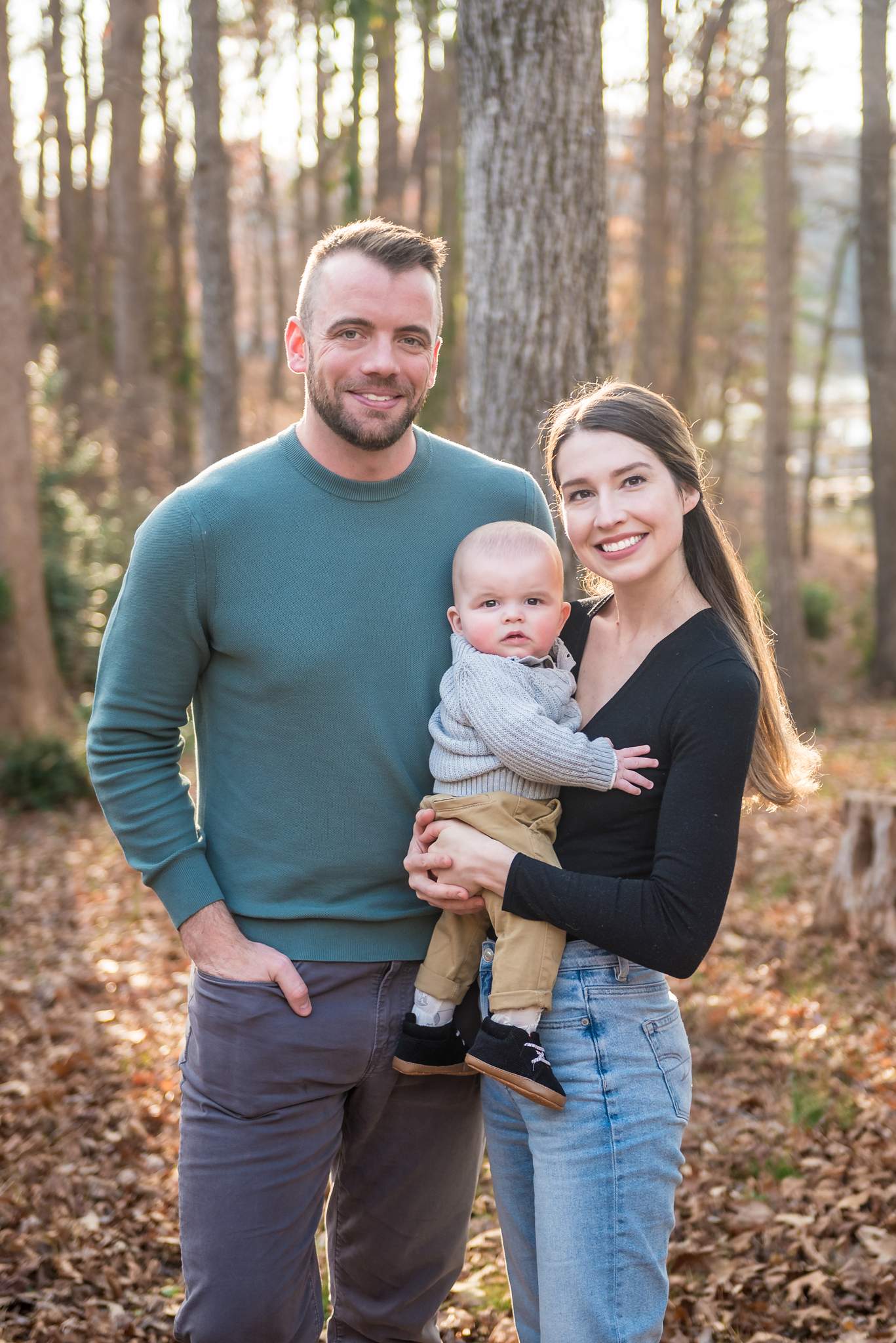 Teal sweater, black top and light jeans on parents holding their baby in gray among fall leaves