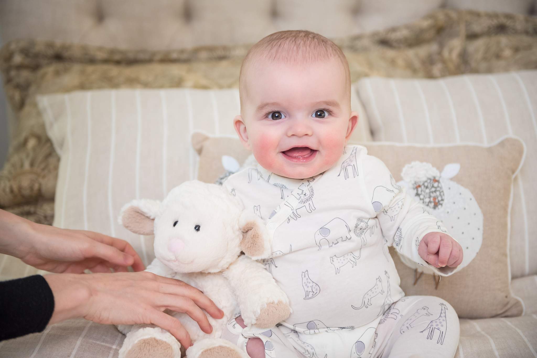 Smiling baby in a white printed sleeper with a plush lamb on a cream settee