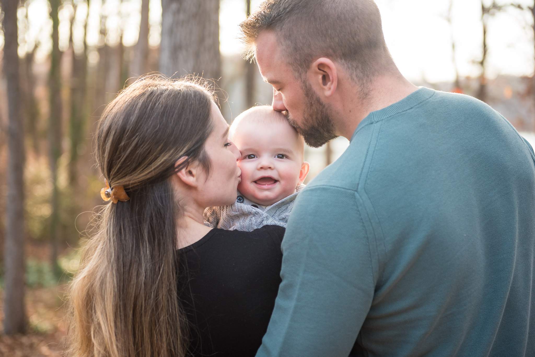 Mom in a black top and dad in a teal sweater kissing their smiling baby in fall light