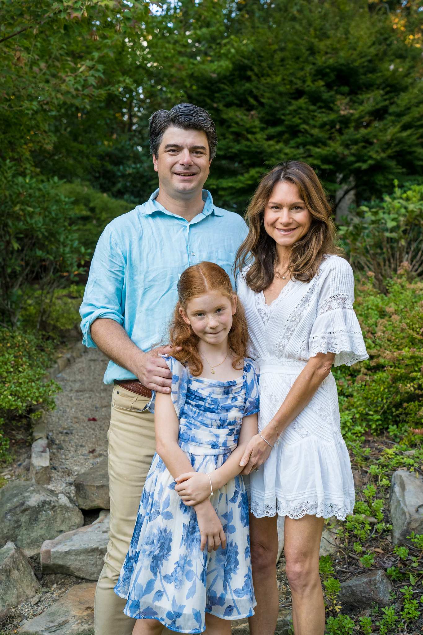 Light blue shirt, white eyelet dress and blue floral dress on a family of three in a garden