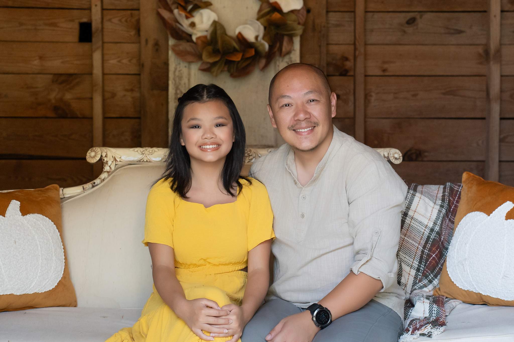 Dad in a beige band-collar shirt beside his daughter in a mustard yellow dress on a white settee