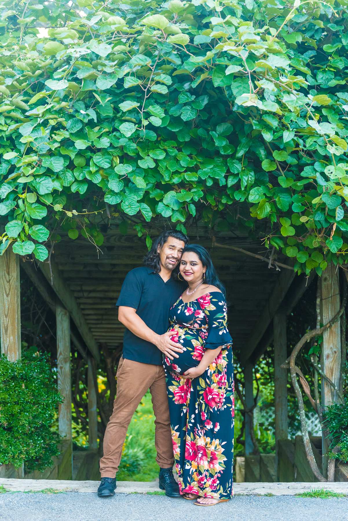 Couple under a vine-covered pergola, mom in a navy floral maxi dress and partner in navy and tan