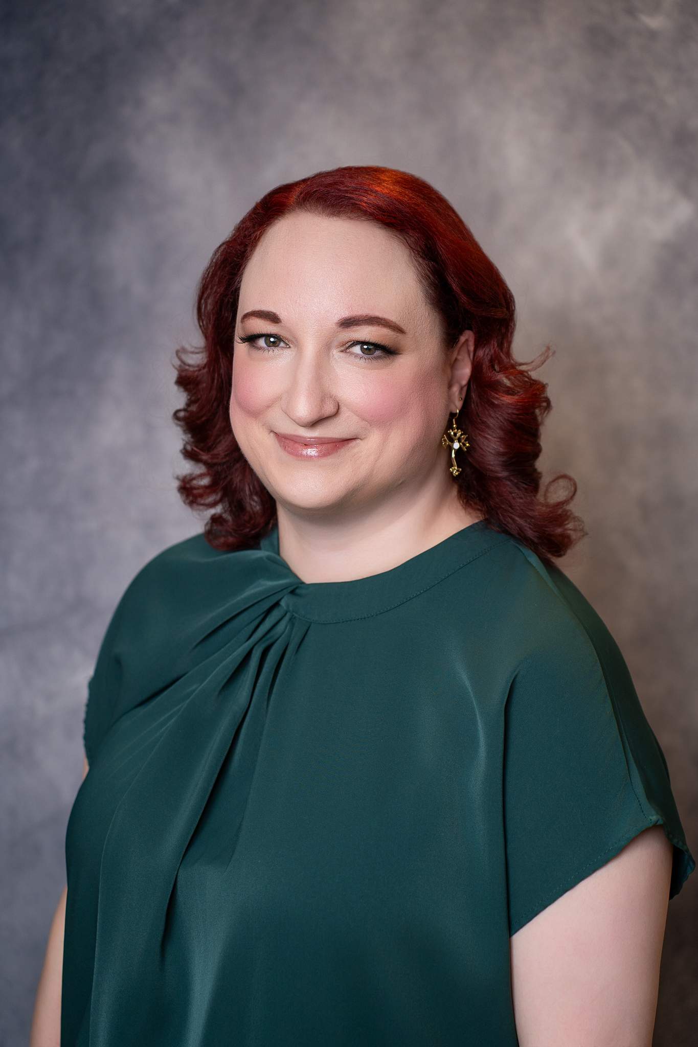 Studio headshot of a smiling woman with red curls in an emerald green top on a gray backdrop