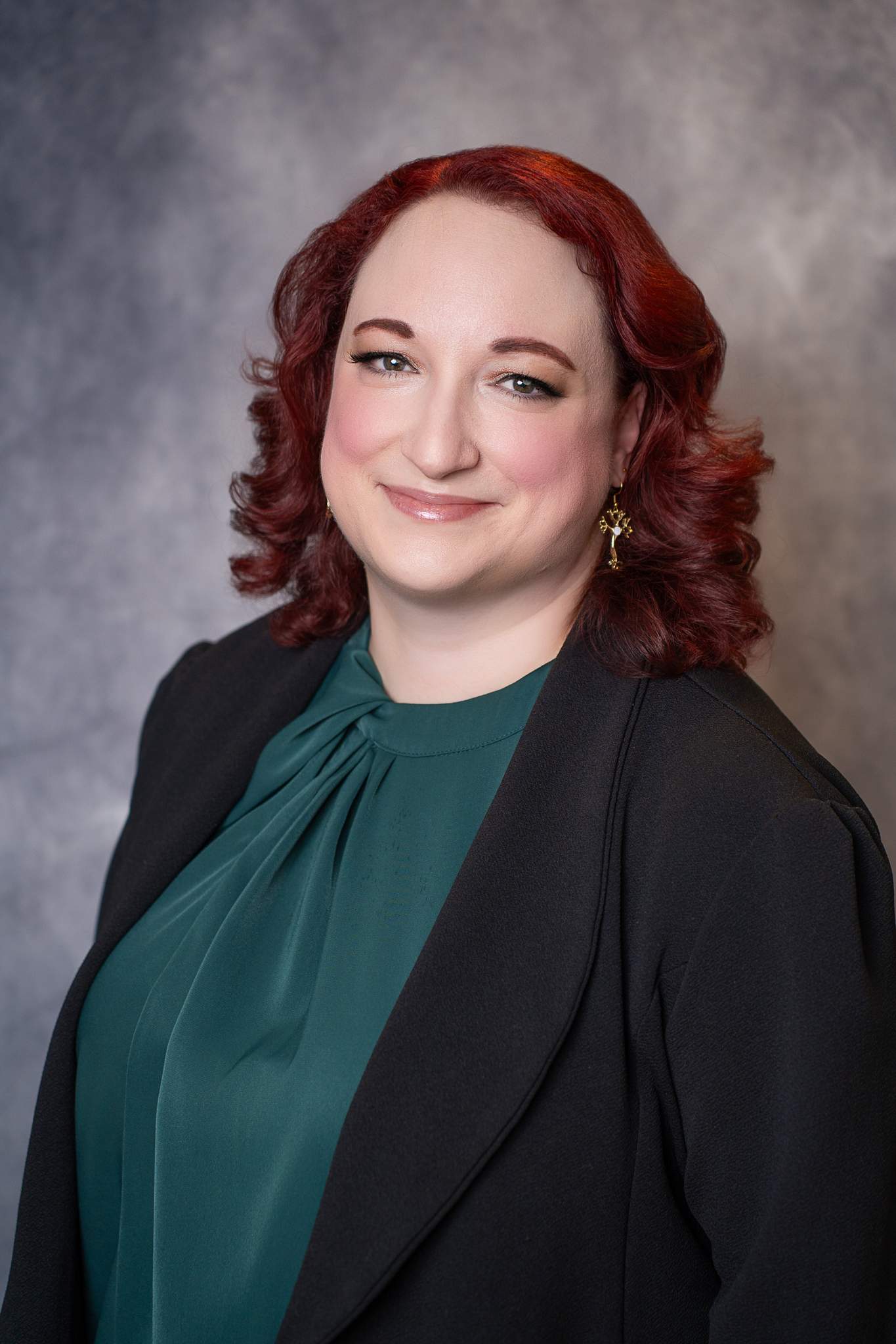 Headshot of a smiling woman with red curls in an emerald top layered under a black blazer on a gray backdrop