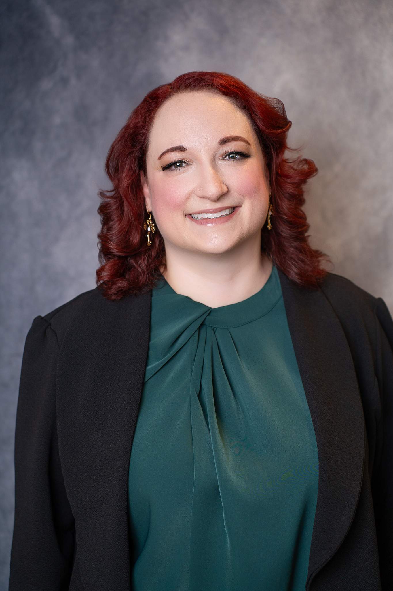 Close headshot of a woman with red curls in an emerald top and black blazer on a gray backdrop