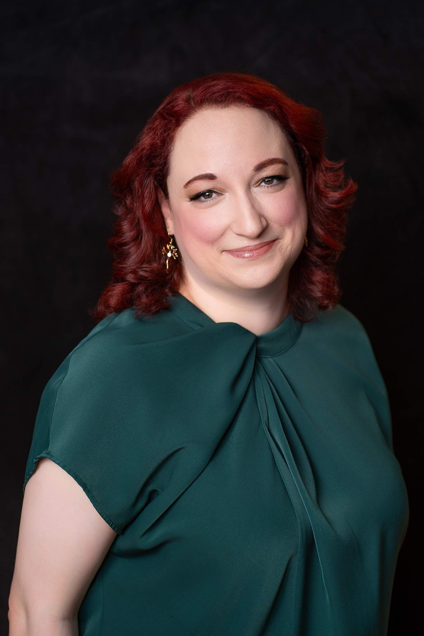 Smiling woman with red curls in an emerald green top on a black studio backdrop