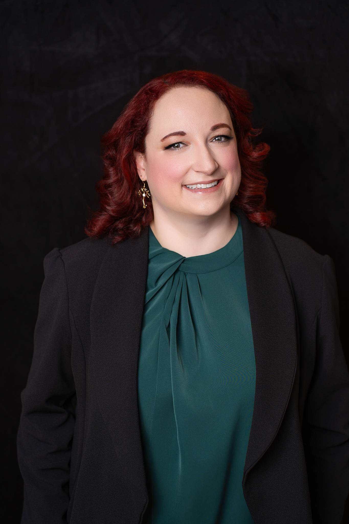 Woman with red curls in an emerald top and black blazer on a black studio backdrop