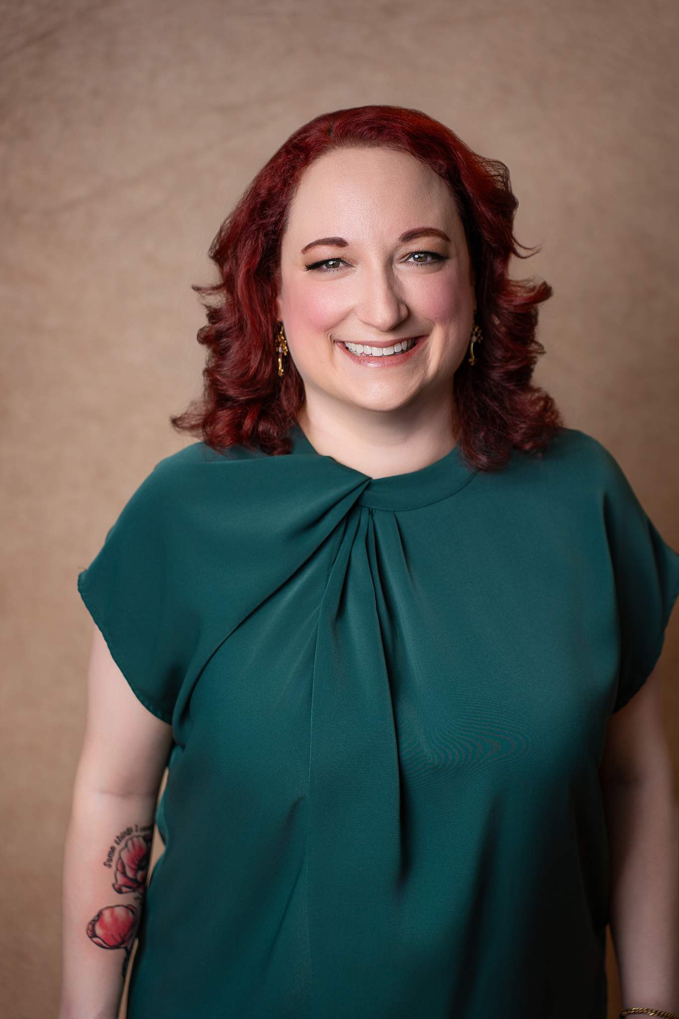 Headshot of a smiling woman with red curls in an emerald green top on a warm tan backdrop