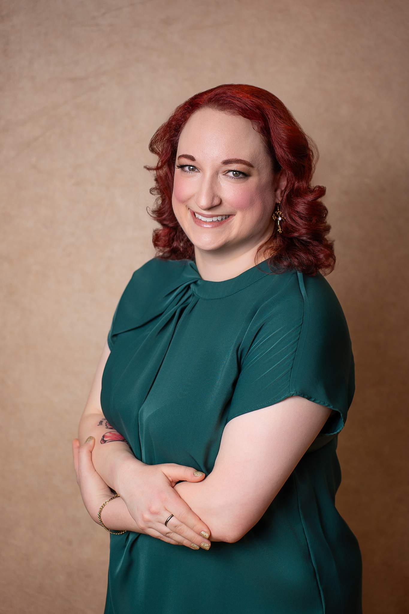 Woman with red curls in an emerald top with arms crossed on a warm tan backdrop