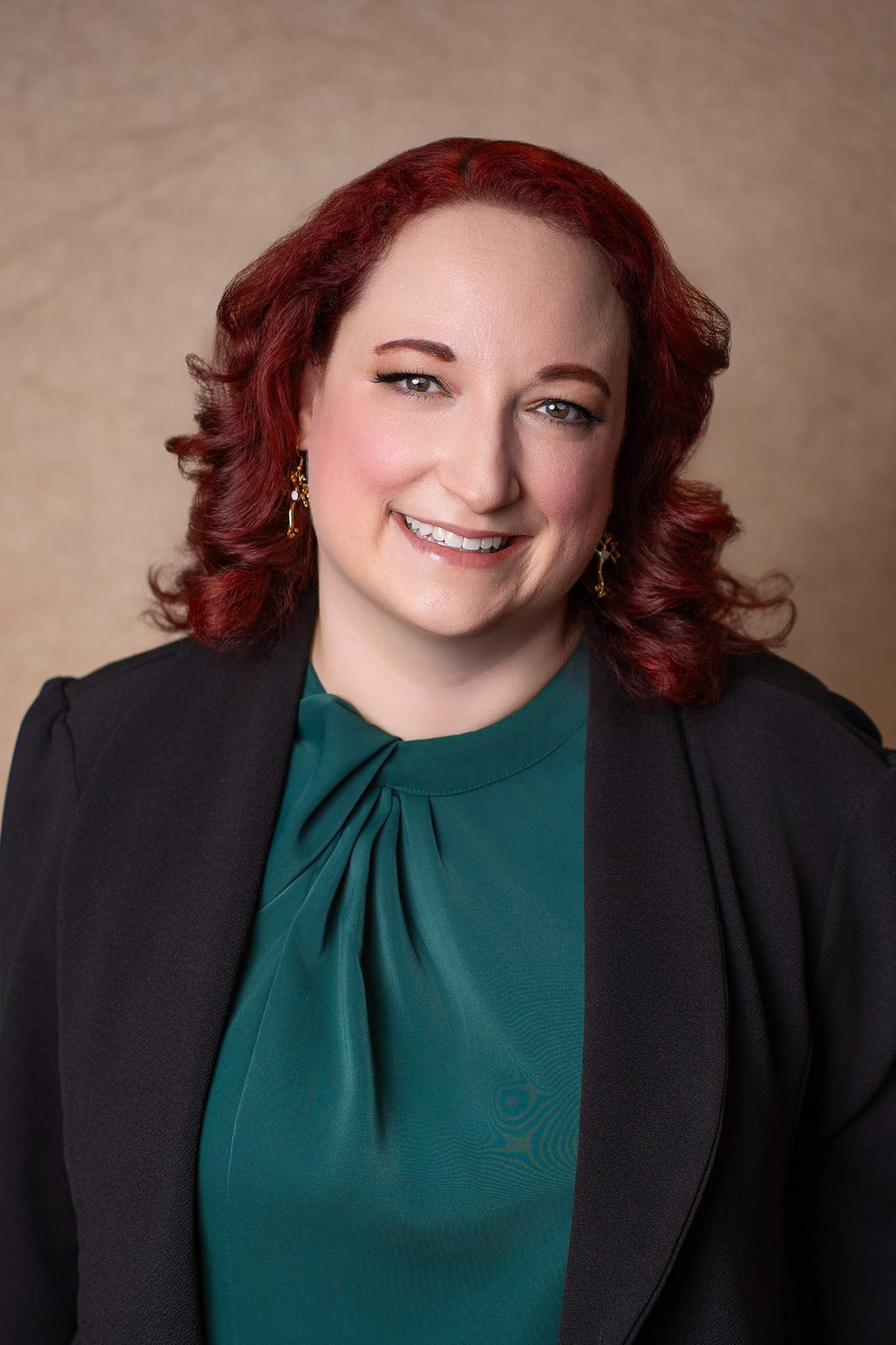 Headshot of a smiling woman with red curls in an emerald top and black blazer on a tan backdrop