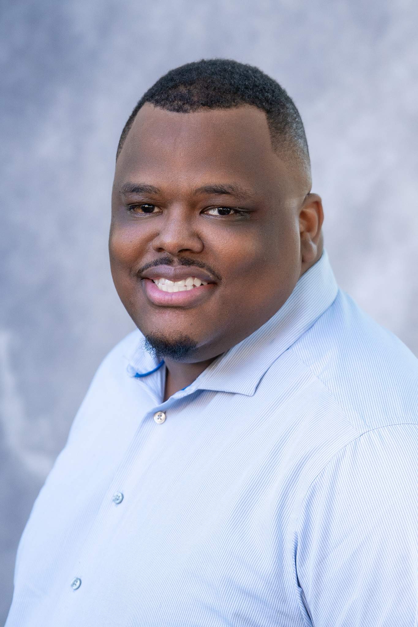 Close headshot of a smiling man in a light blue button-down shirt on a blue studio backdrop