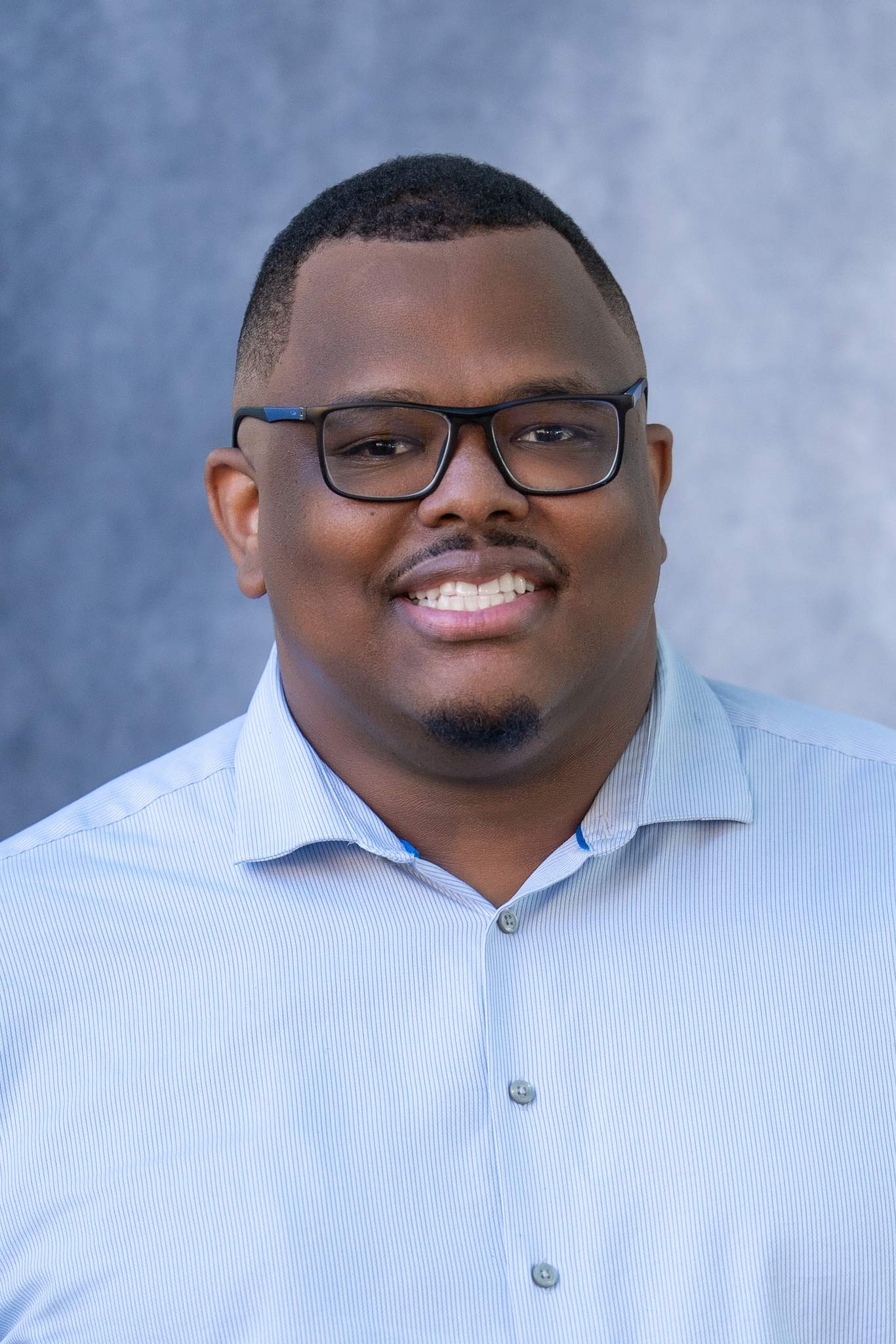Close headshot of a smiling man with glasses in a light blue shirt on a blue studio backdrop