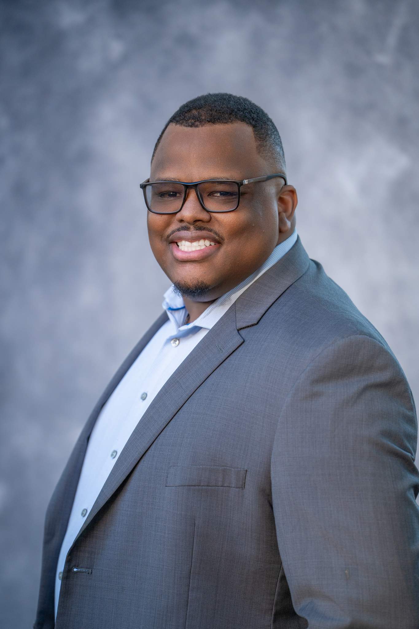 Angled business headshot of a man with glasses in a gray jacket and light blue shirt