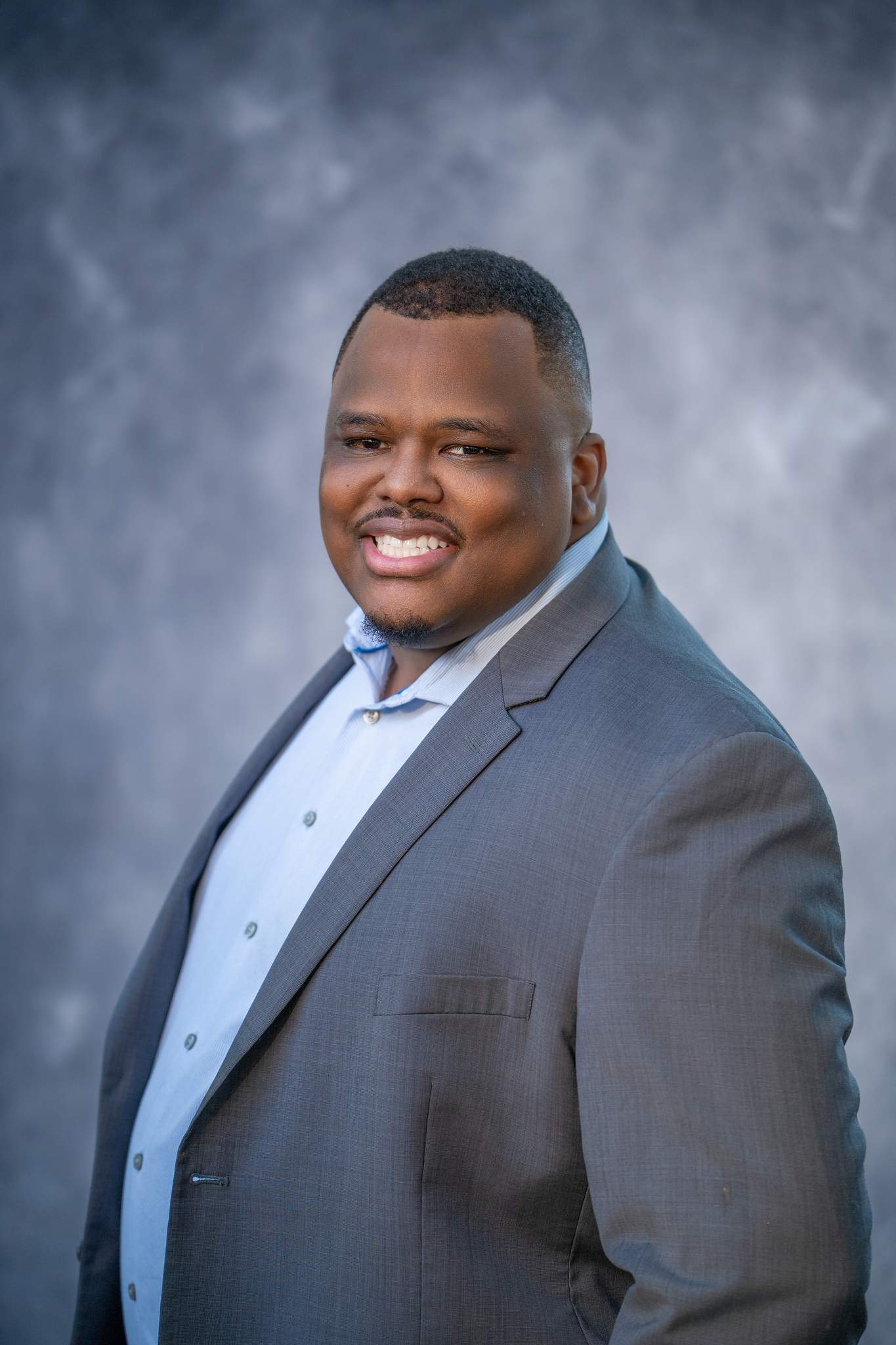 Smiling man in a gray suit jacket and light blue shirt on a blue studio backdrop