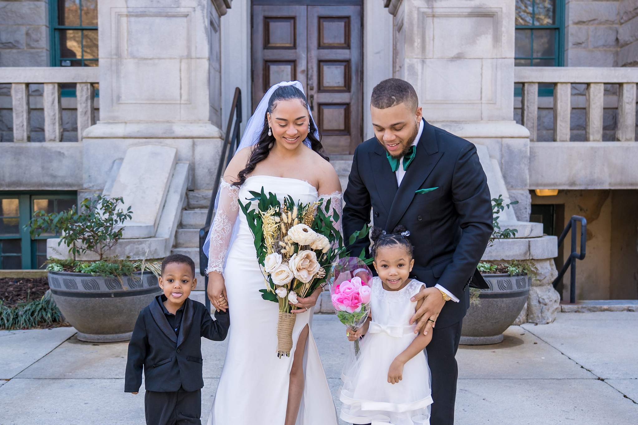 Bride in strapless gown with lace sleeves, groom in black suit, and two dressed-up children on stone steps