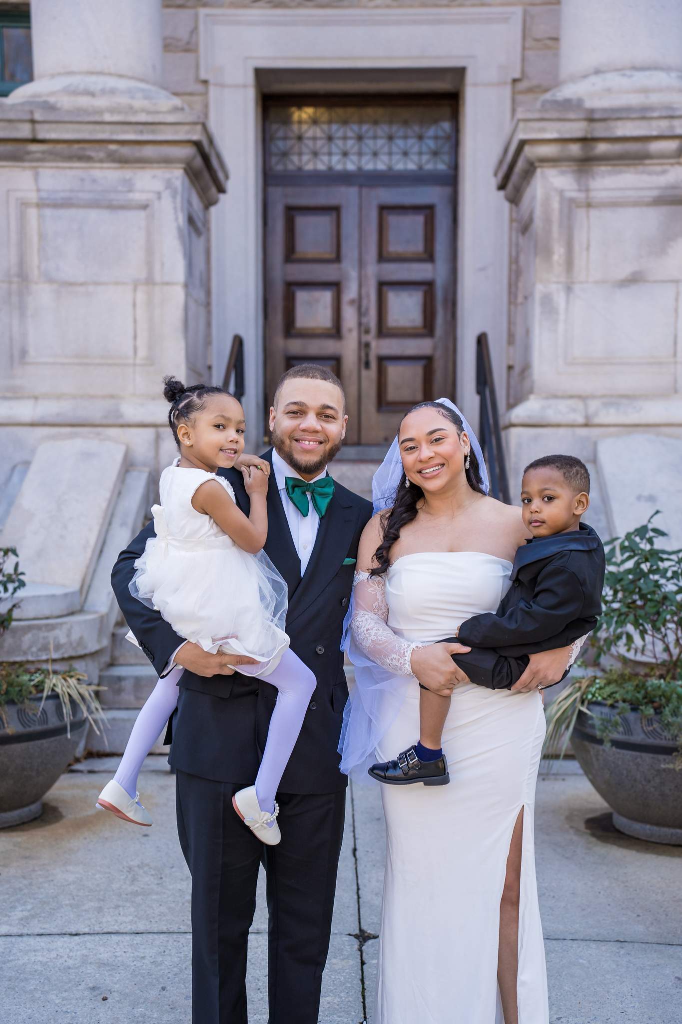 Couple in a black suit and a white slit gown, each holding a dressed-up child on stone steps