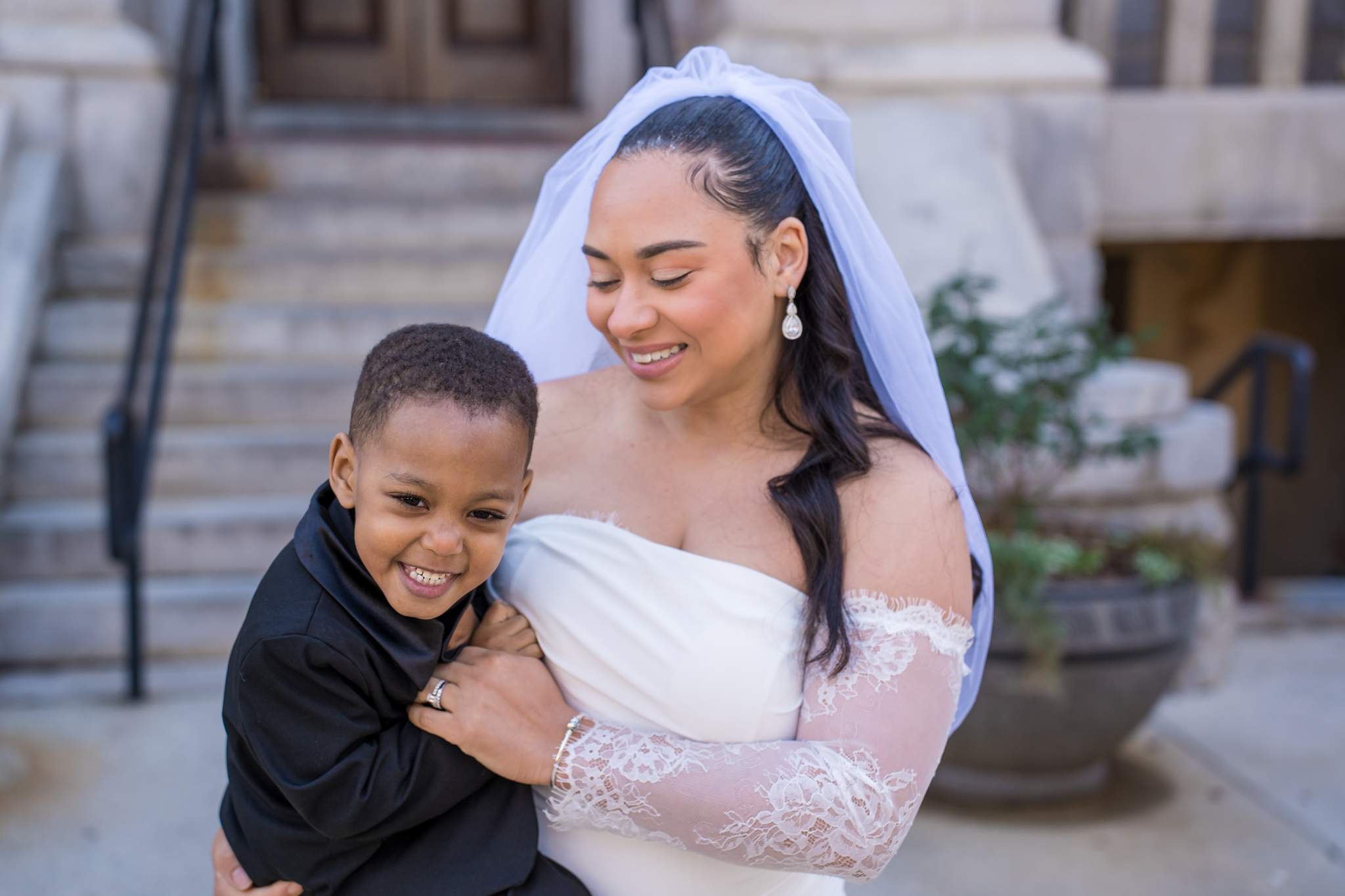 Bride in sheer lace sleeves and a tulle veil hugging a smiling boy in a black suit