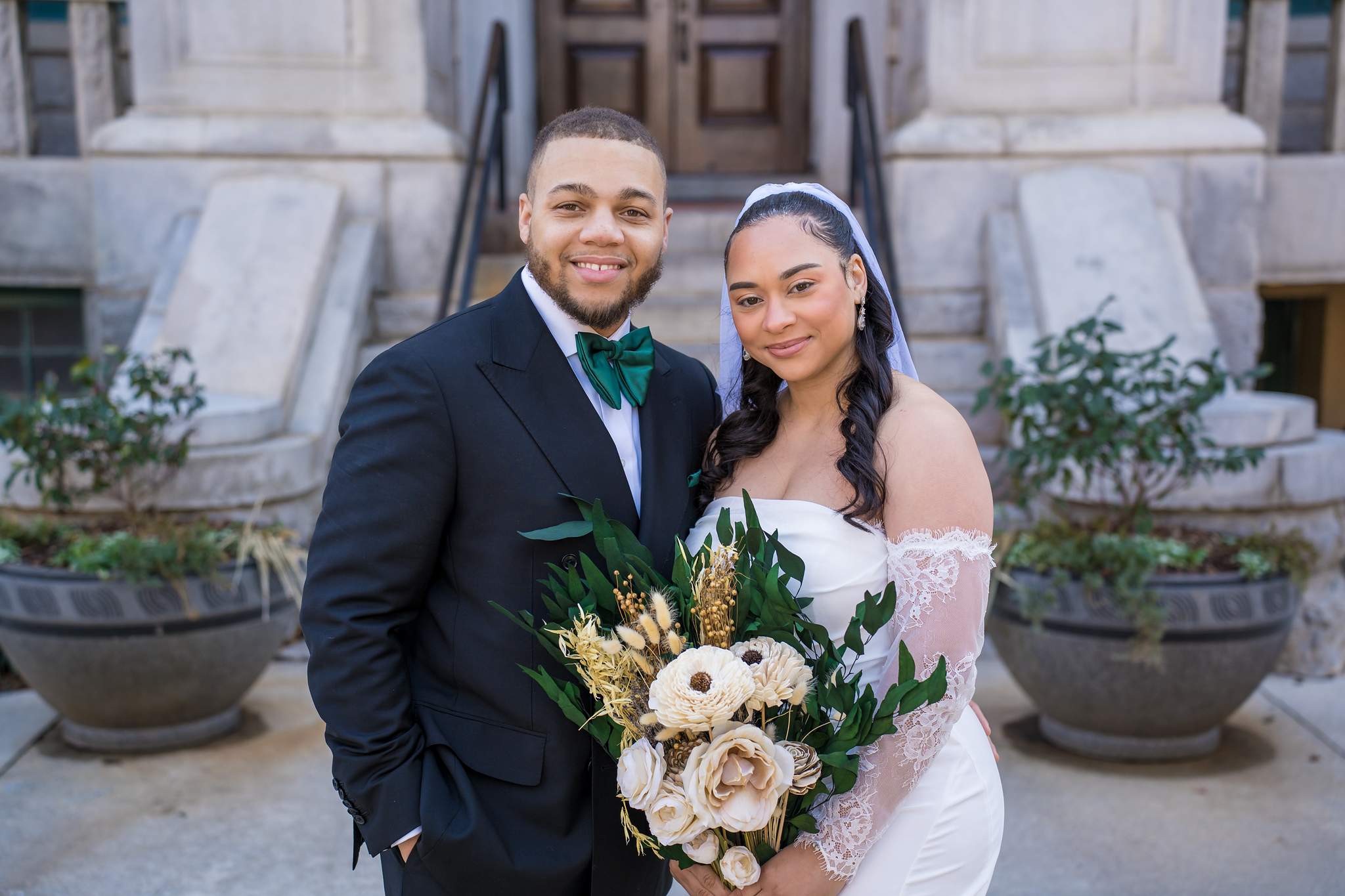 Groom in a black suit with an emerald bow tie beside the bride holding a cream dried-flower bouquet