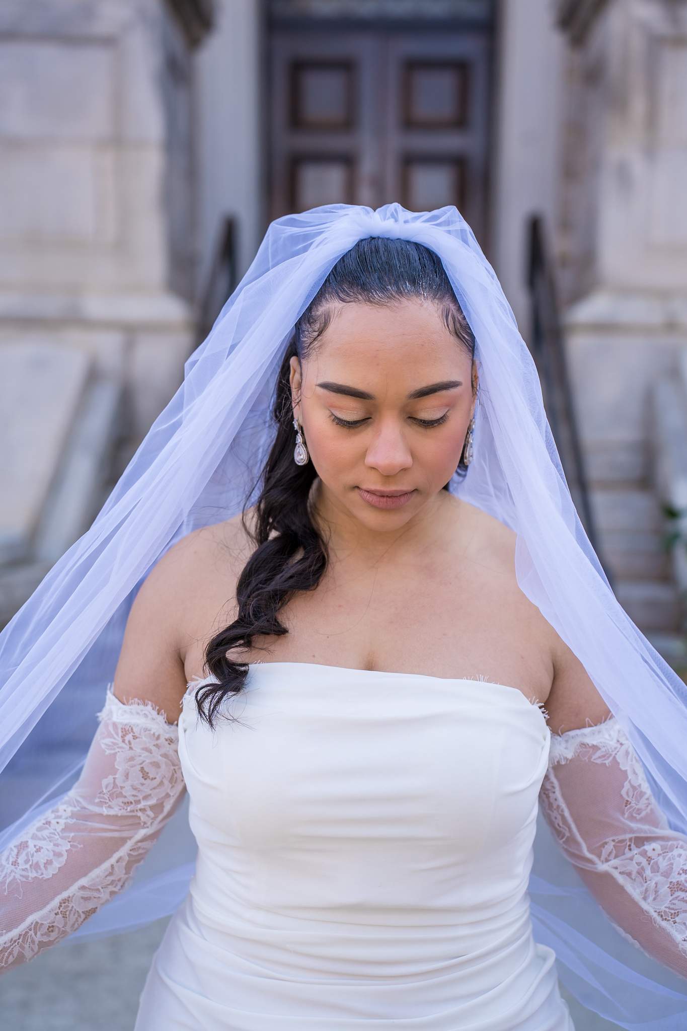 Bride in a strapless white gown, sheer lace sleeves, drop earrings and a long tulle veil