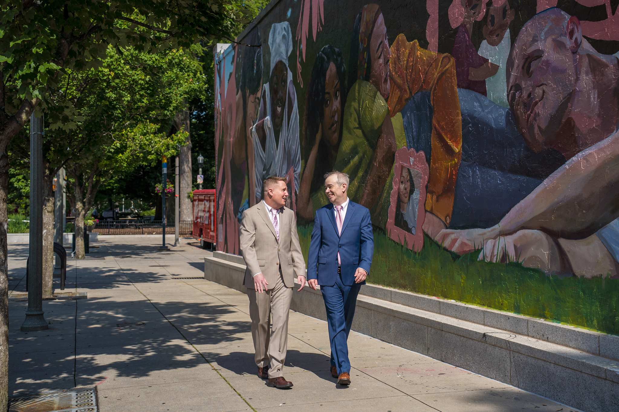 Two grooms in a light tan suit and a blue suit walking past a mural in downtown Decatur