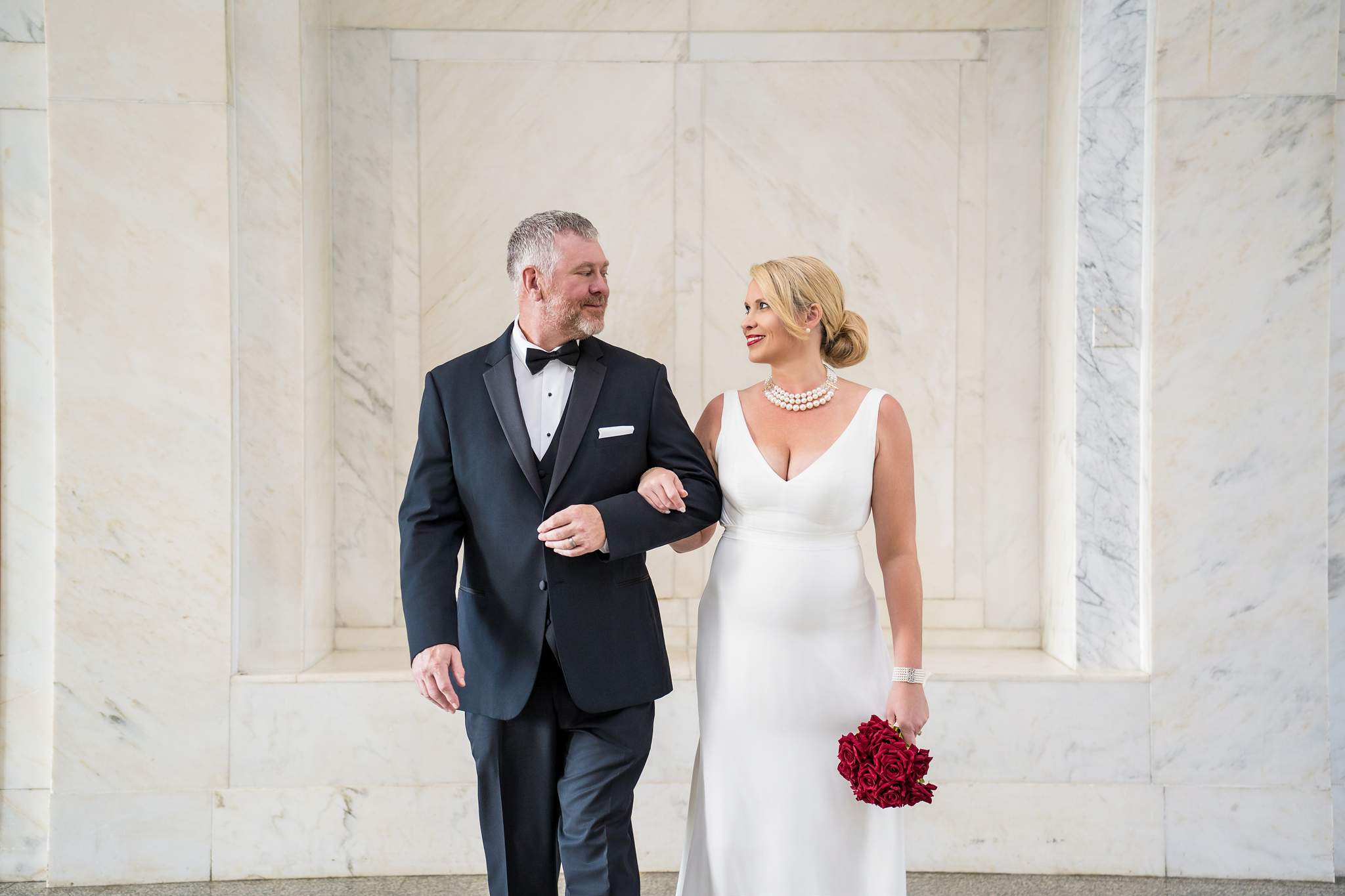 Sleek white sheath gown and black tuxedo as the couple walks arm in arm through a marble hall