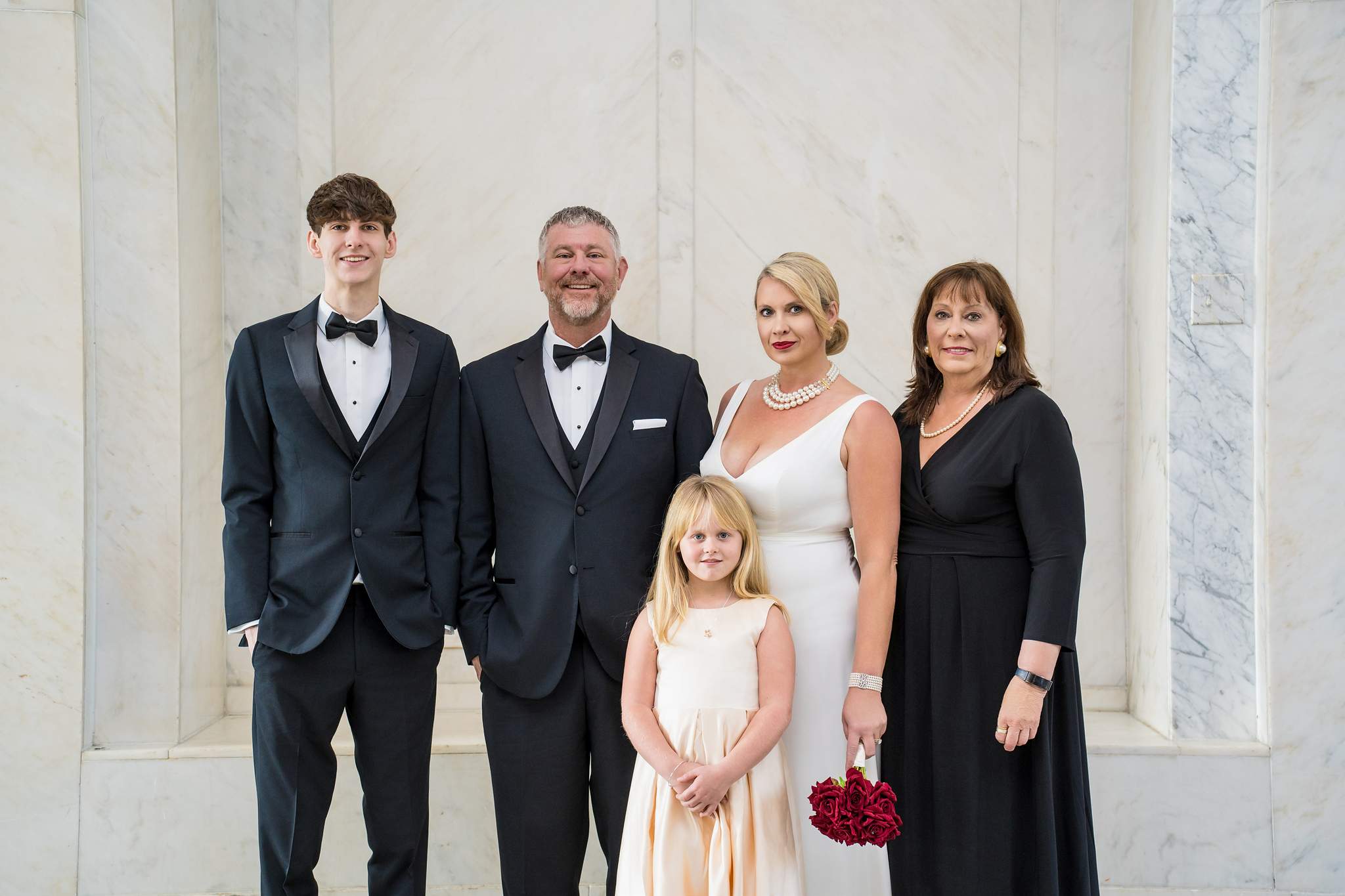 Formal courthouse wedding attire in a marble hall: black tuxedos, white sheath gown, black dress and champagne satin
