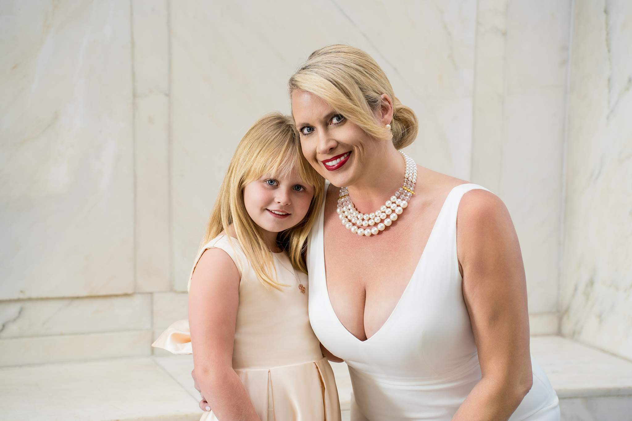 Bride in a white V-neck gown and pearls with a girl in champagne satin against white marble