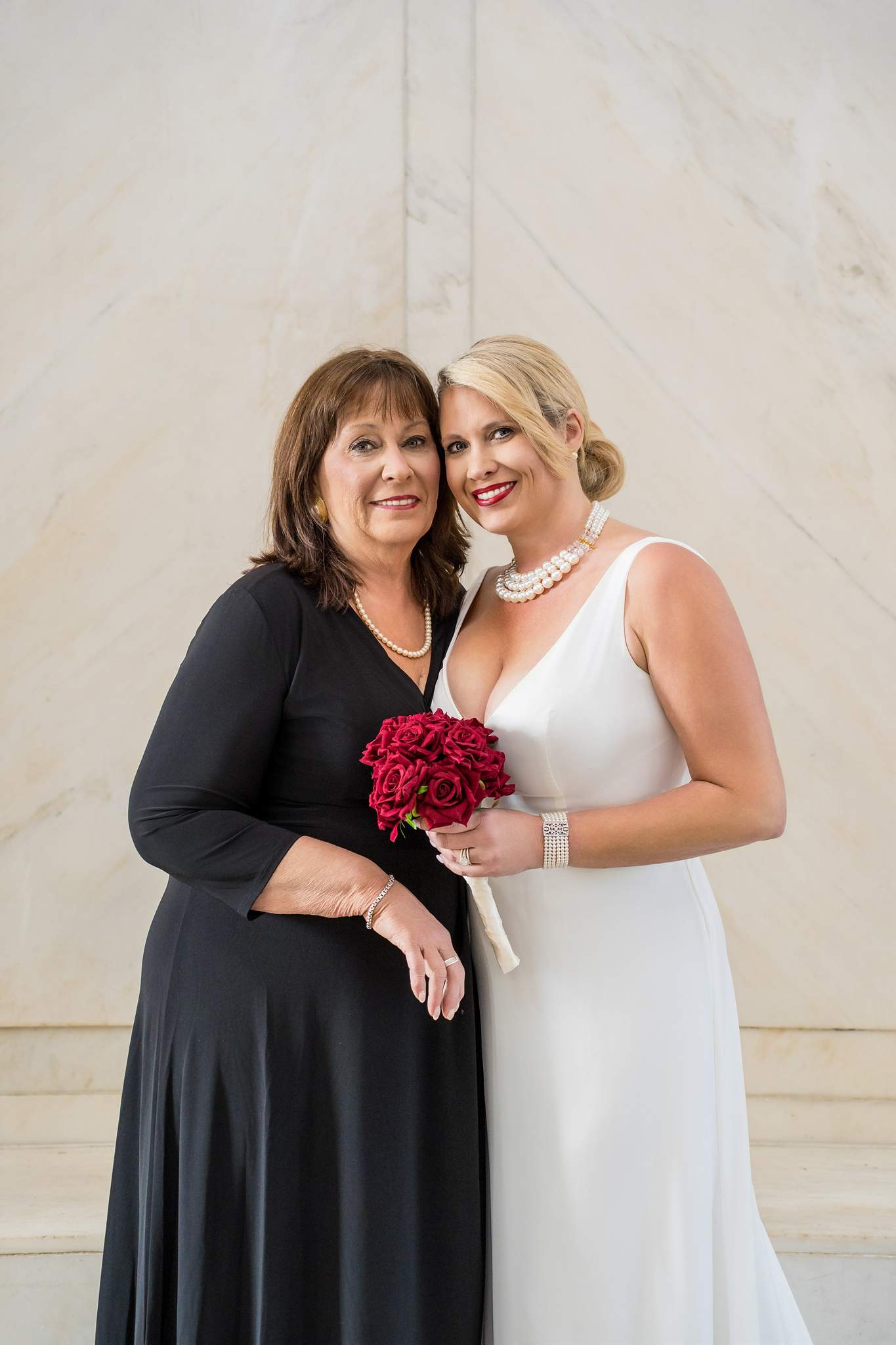 Family member in a long black dress beside the bride in white holding red roses against white marble