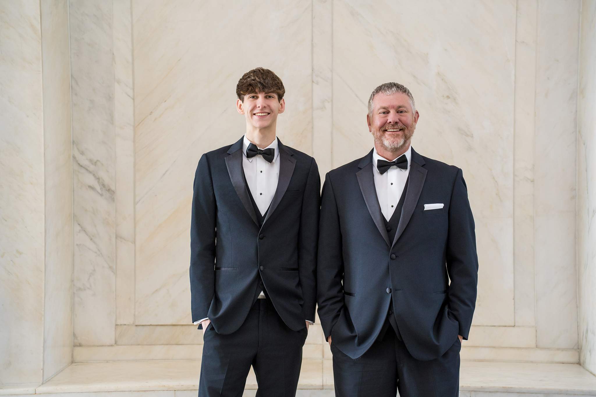 Groom and a teen family member in matching black tuxedos and bow ties against white marble walls