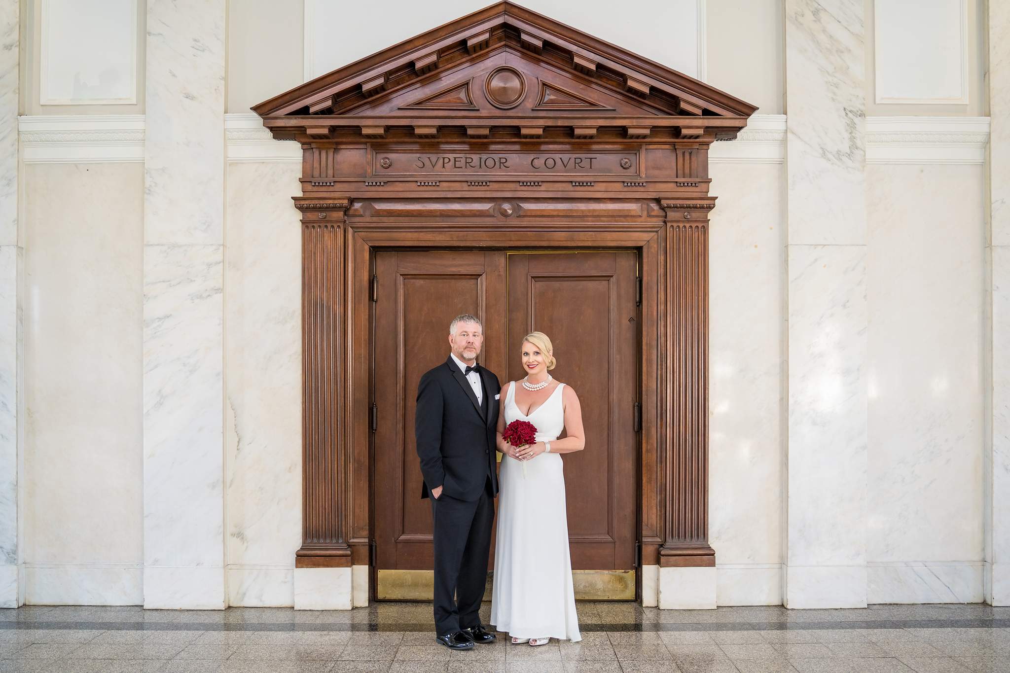 Bride in a white V-neck sheath gown and groom in a black tuxedo at carved courtroom doors